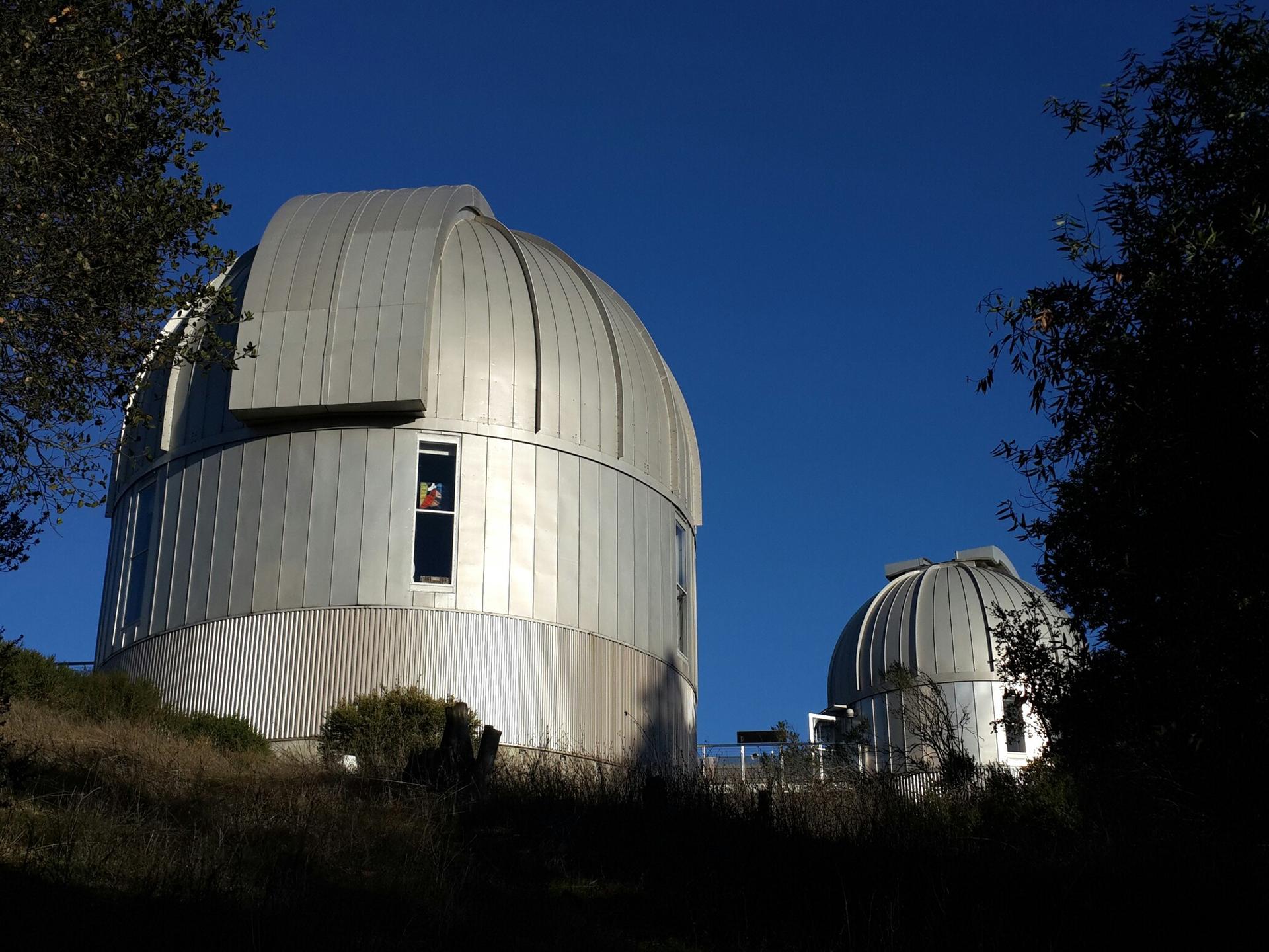 Two large observatory domes stand on a hillside under a clear blue sky.