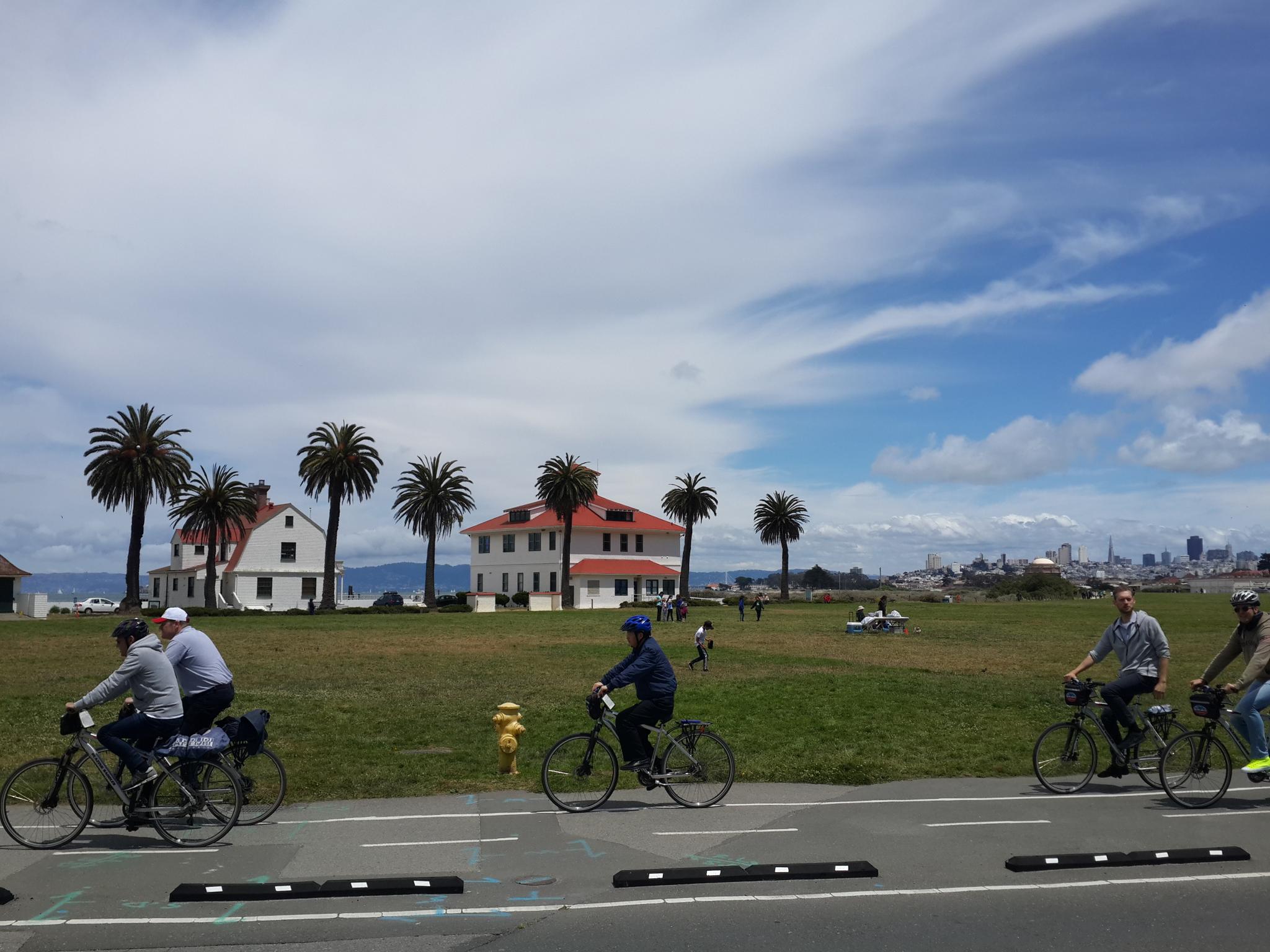 Cyclists ride on a paved path past palm trees and white buildings.