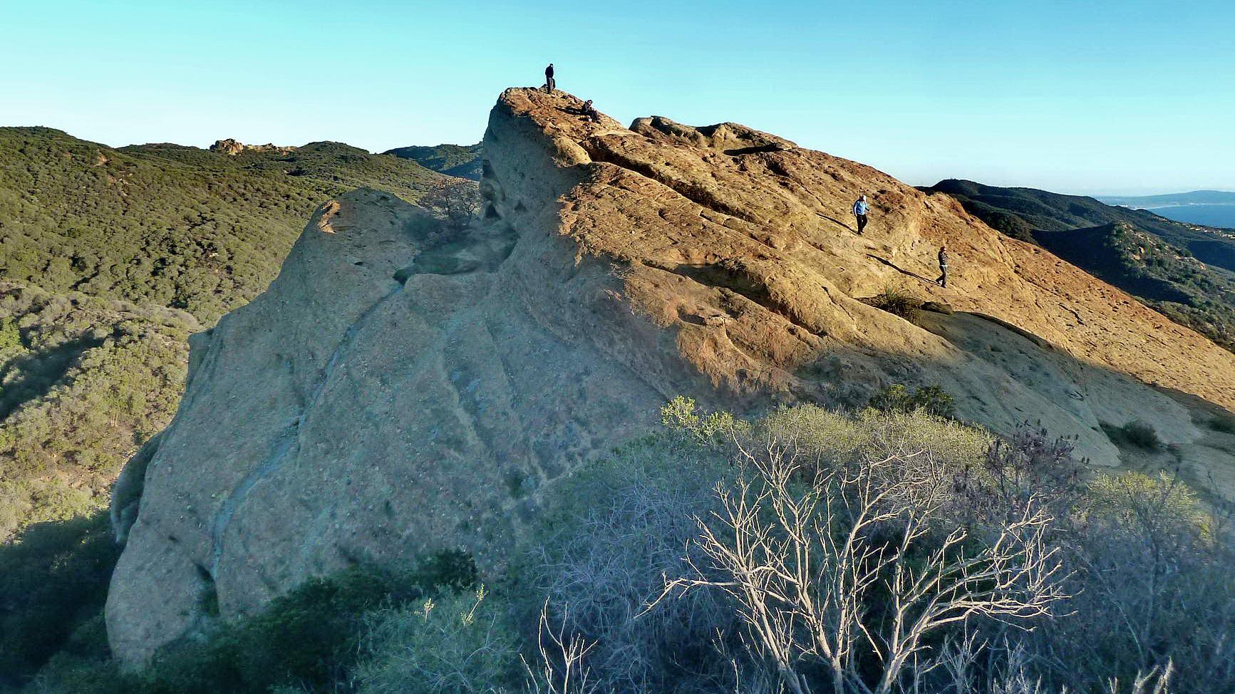 Hikers on a sunlit rocky ridge with green hills in the background.