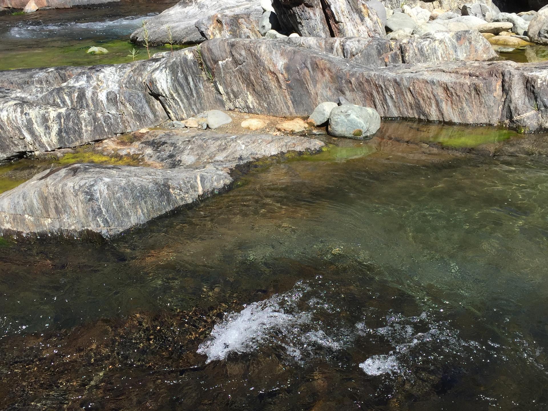 Clear river water flows over and around large, layered rocks in sunlight.