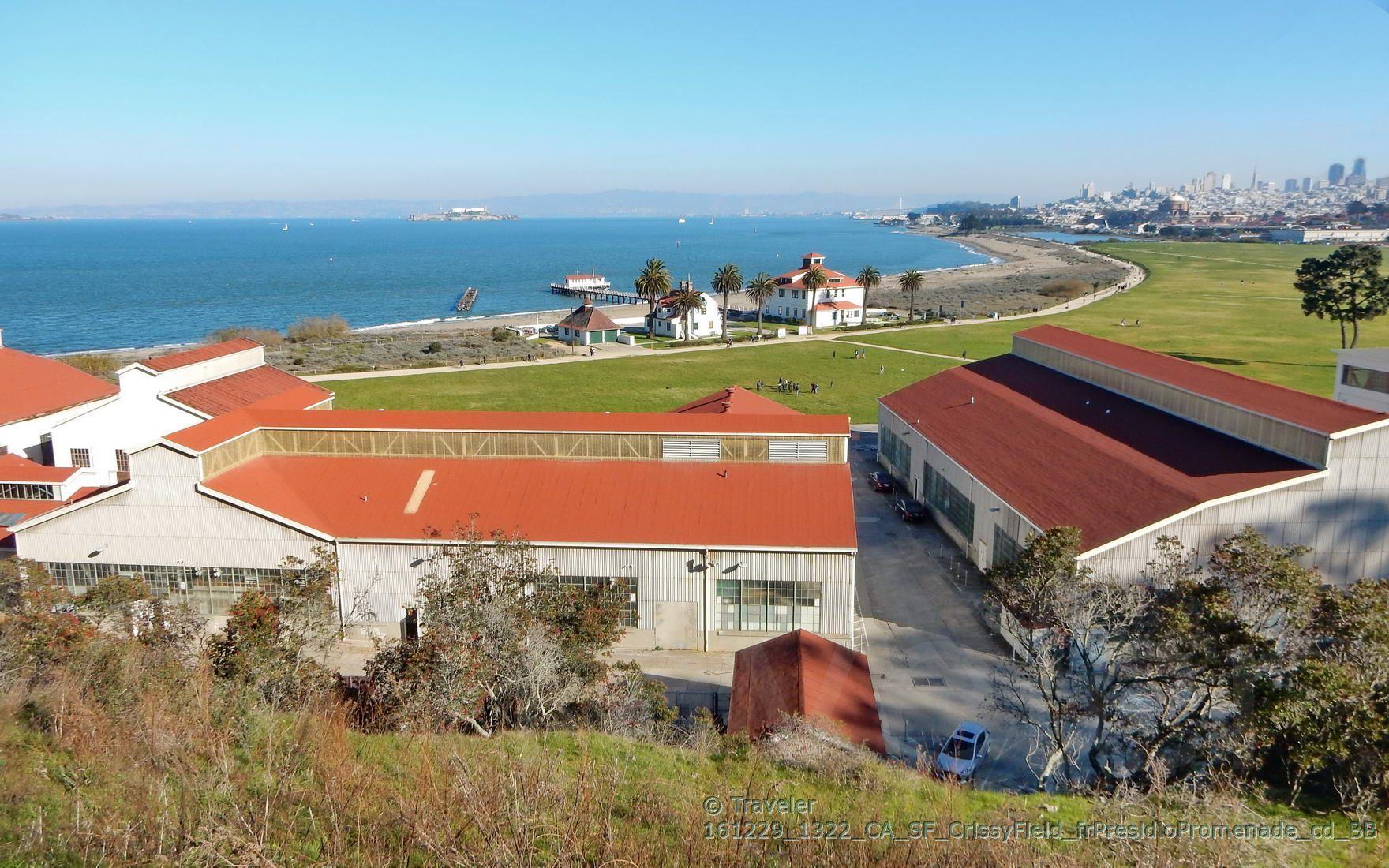 Red-roofed buildings by a bay, green lawn, distant city under clear sky.