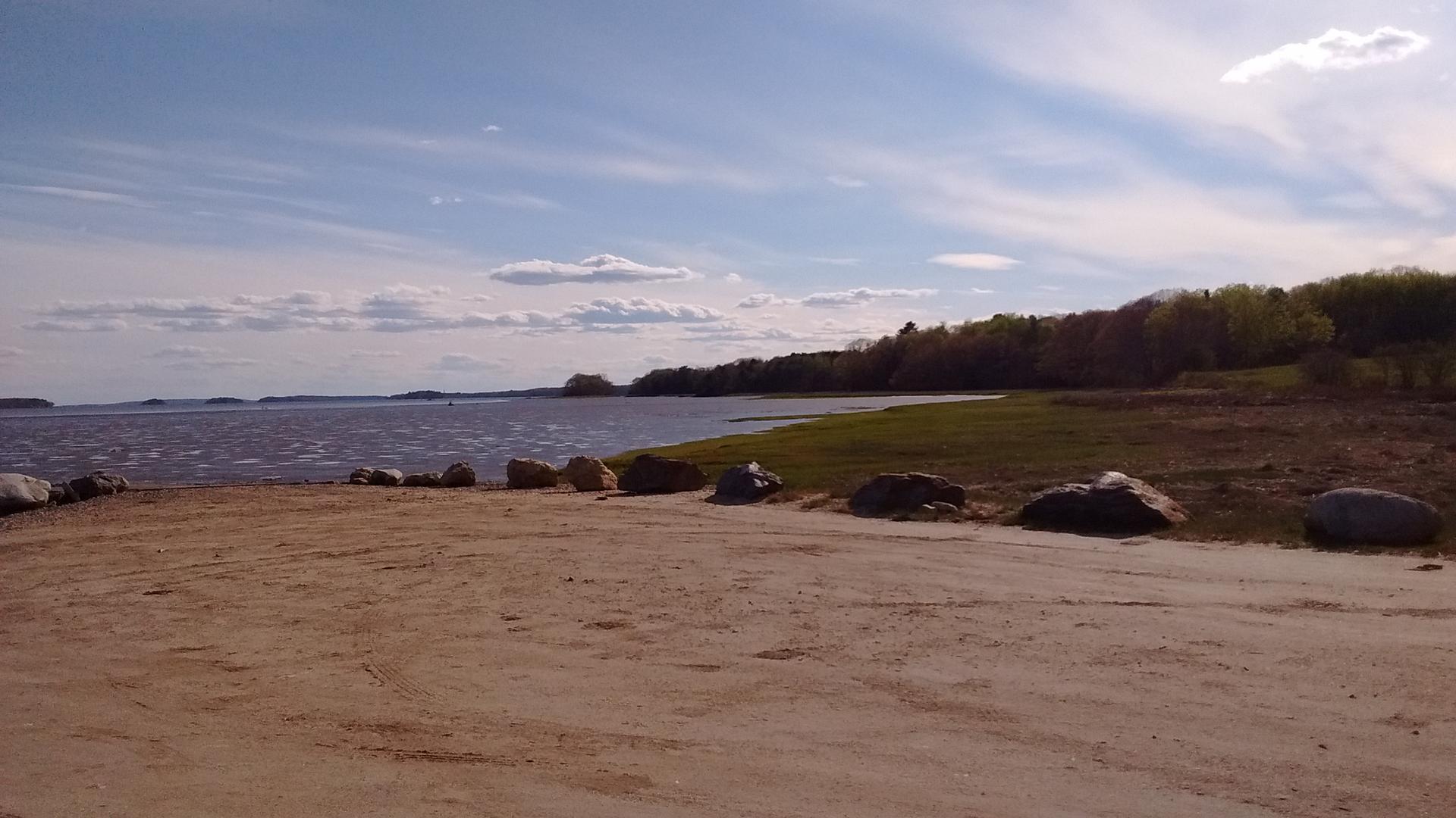 Sandy foreground, calm water, and distant treeline under a bright, cloudy sky.