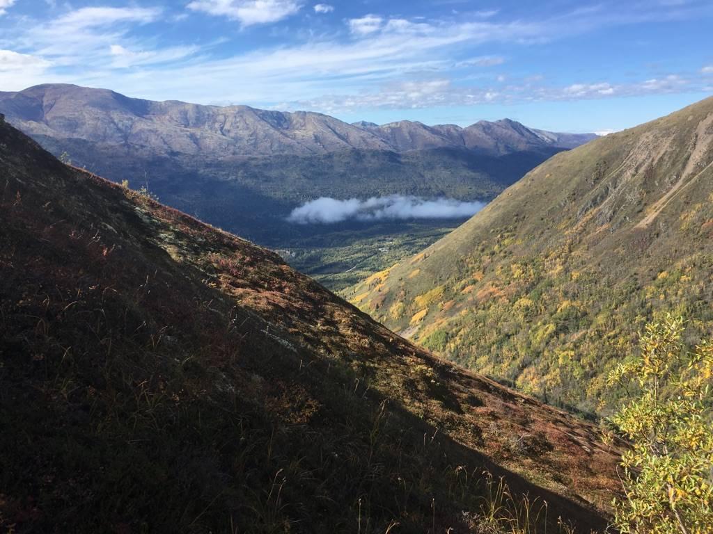 Steep mountain slopes frame a valley with distant clouds under a blue sky.