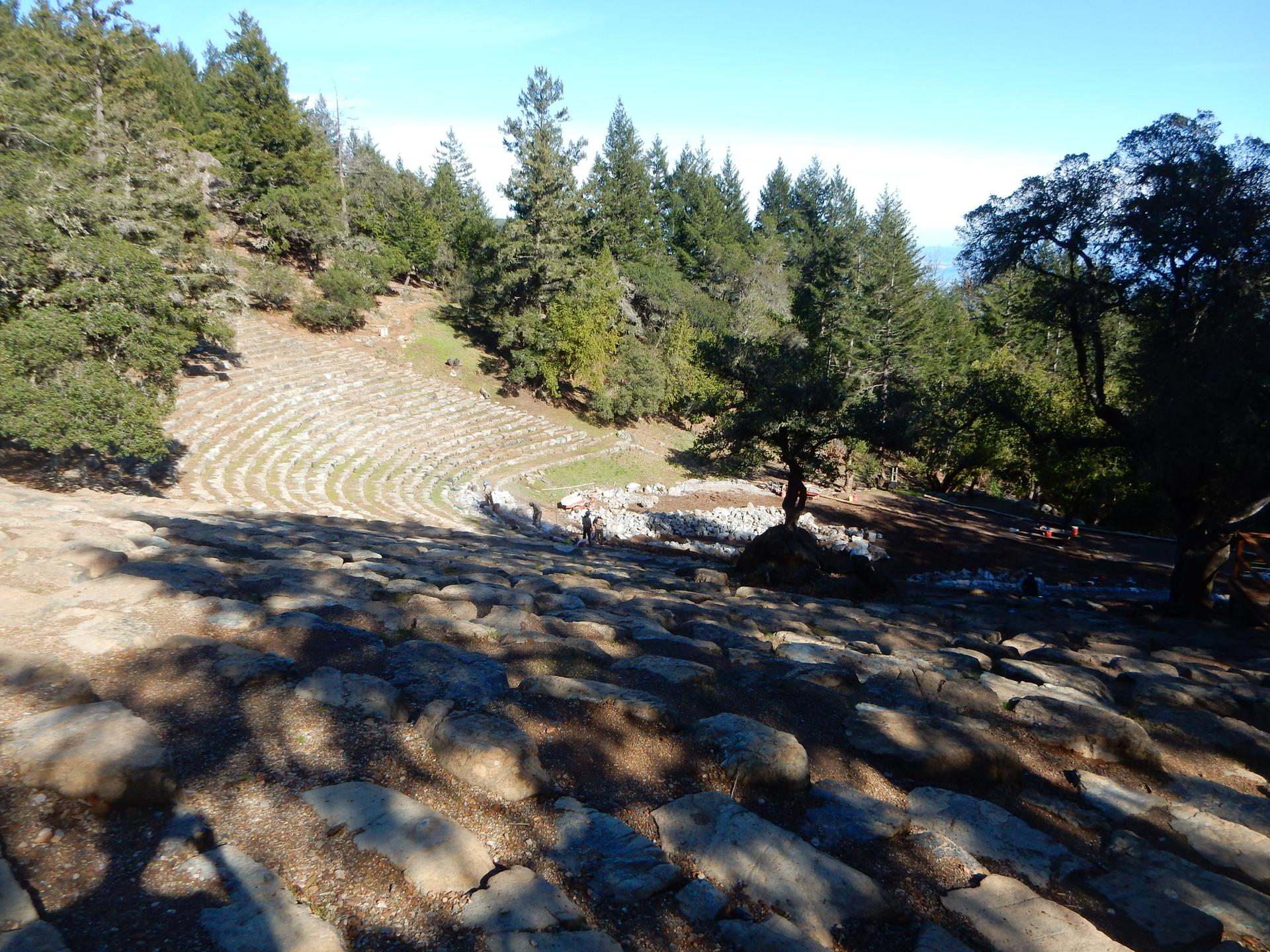 Stone amphitheater carved into a sunny forested hillside with distant figures.