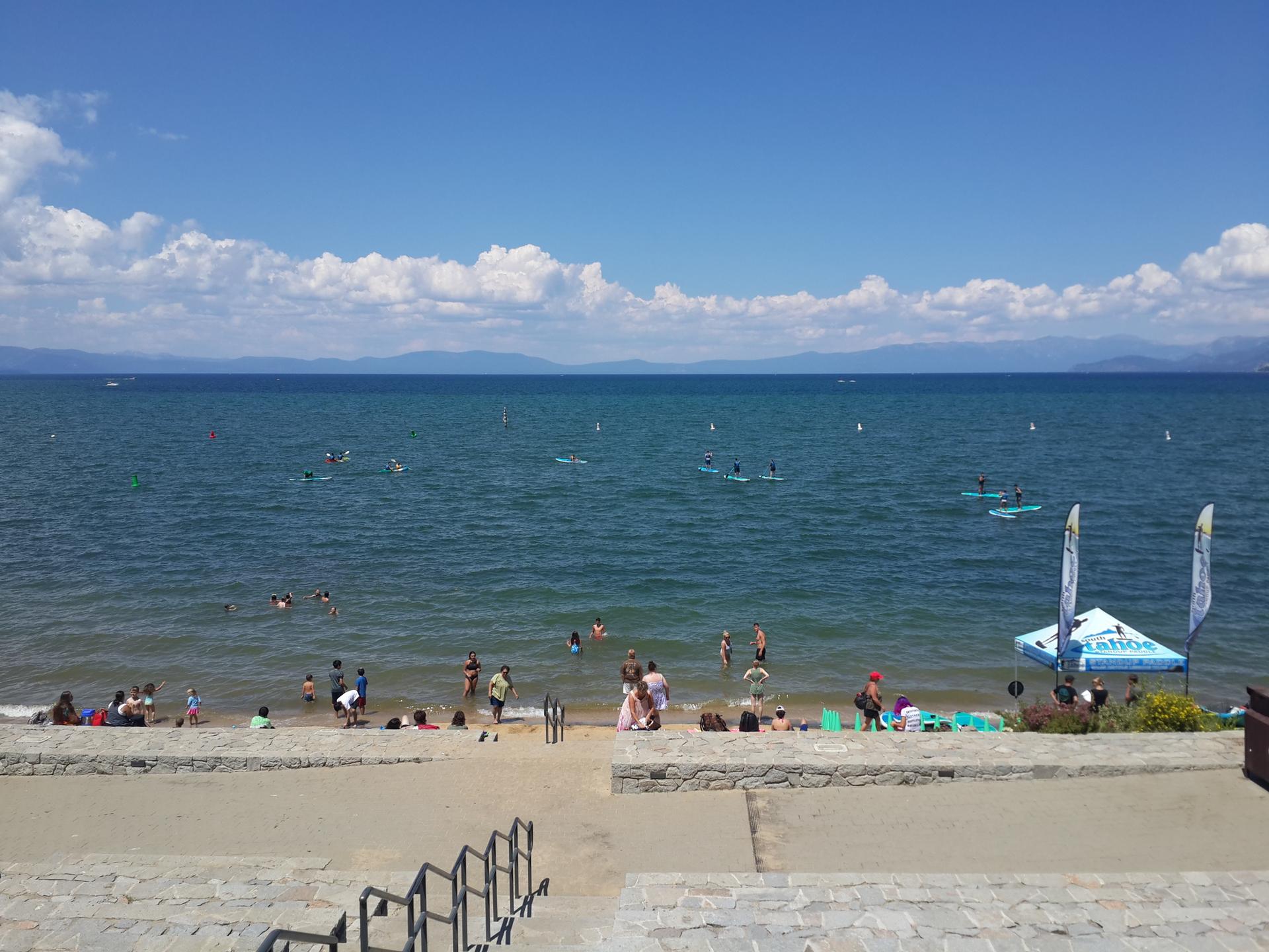 Crowded beach with many people swimming and paddleboarding on a sunny day.