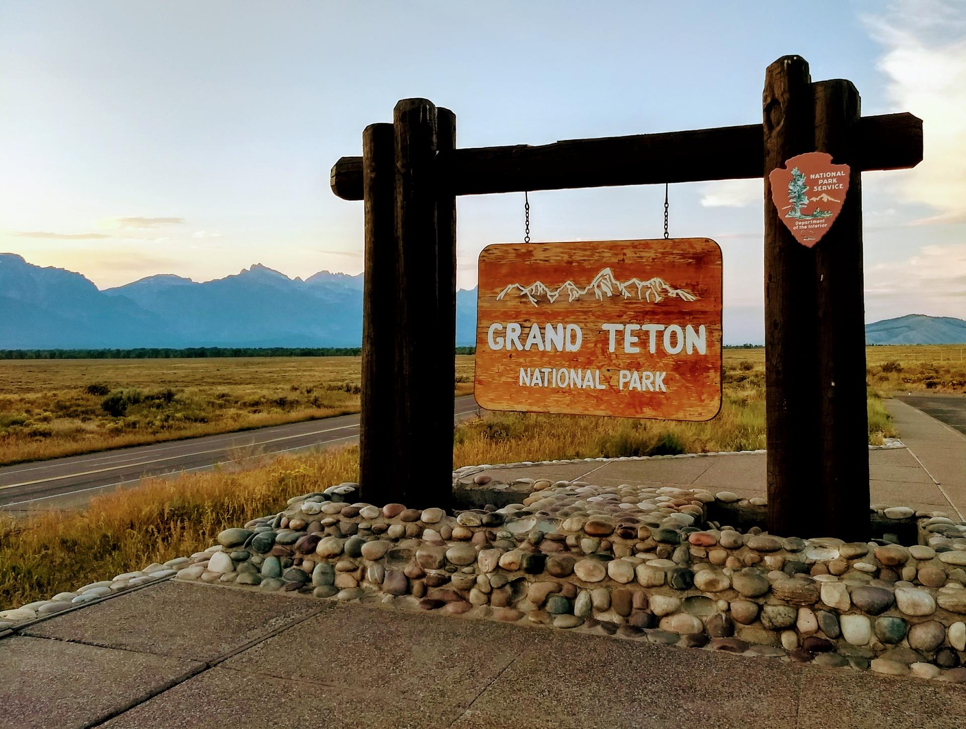 A wooden park entrance sign with mountains and a road at sunset.