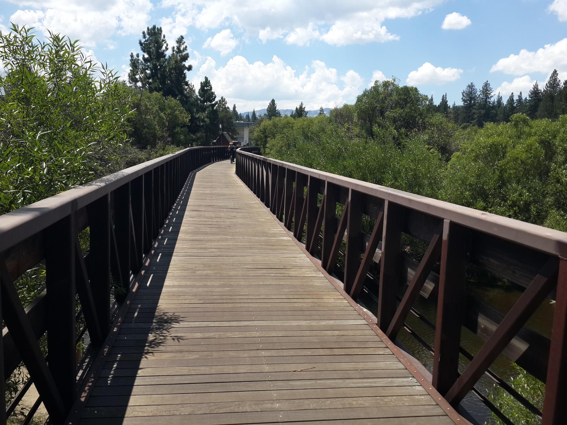 A long wooden bridge curves through green trees under a partly cloudy sky.