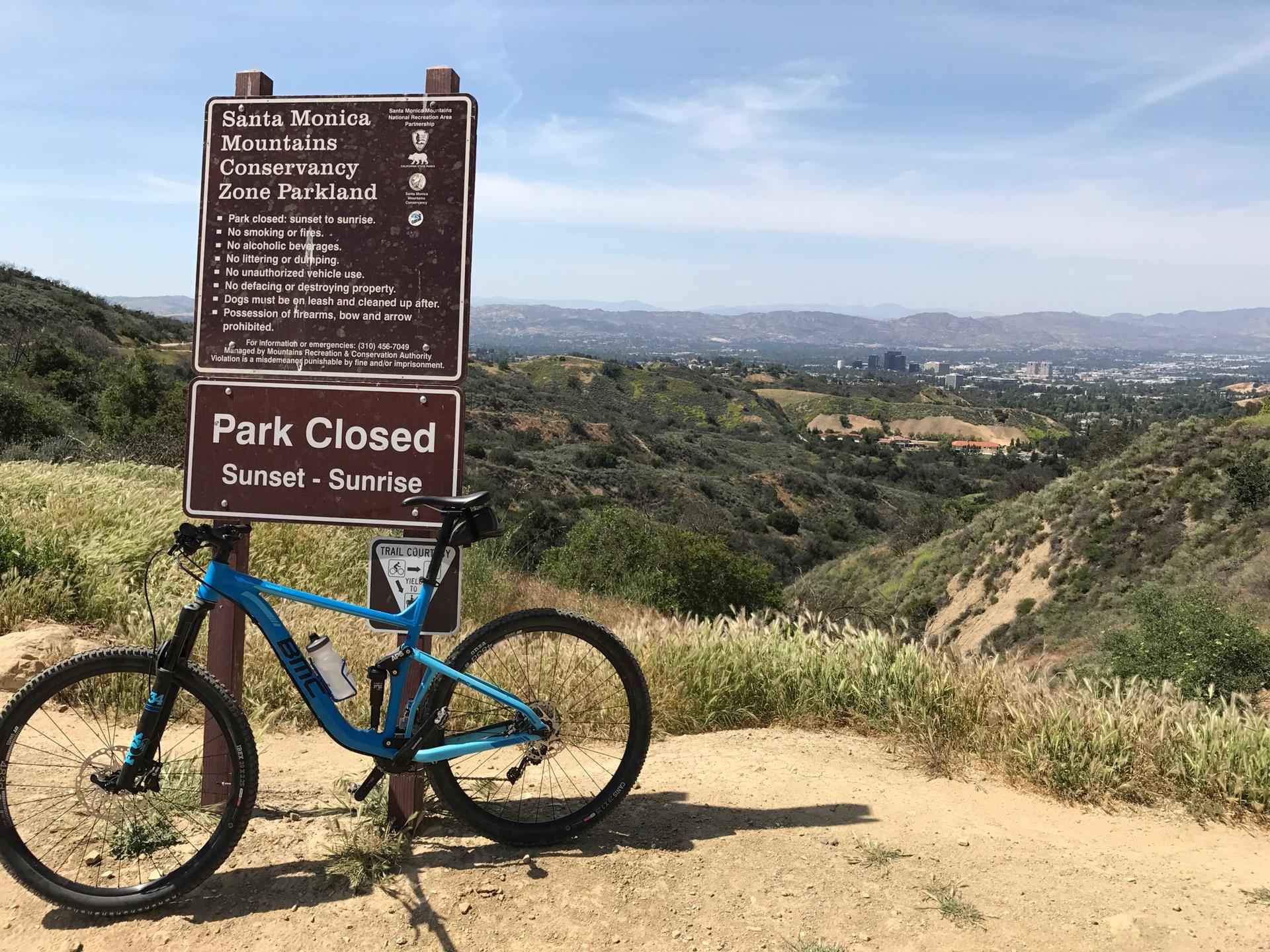 A blue mountain bike rests beside a park sign overlooking a valley.
