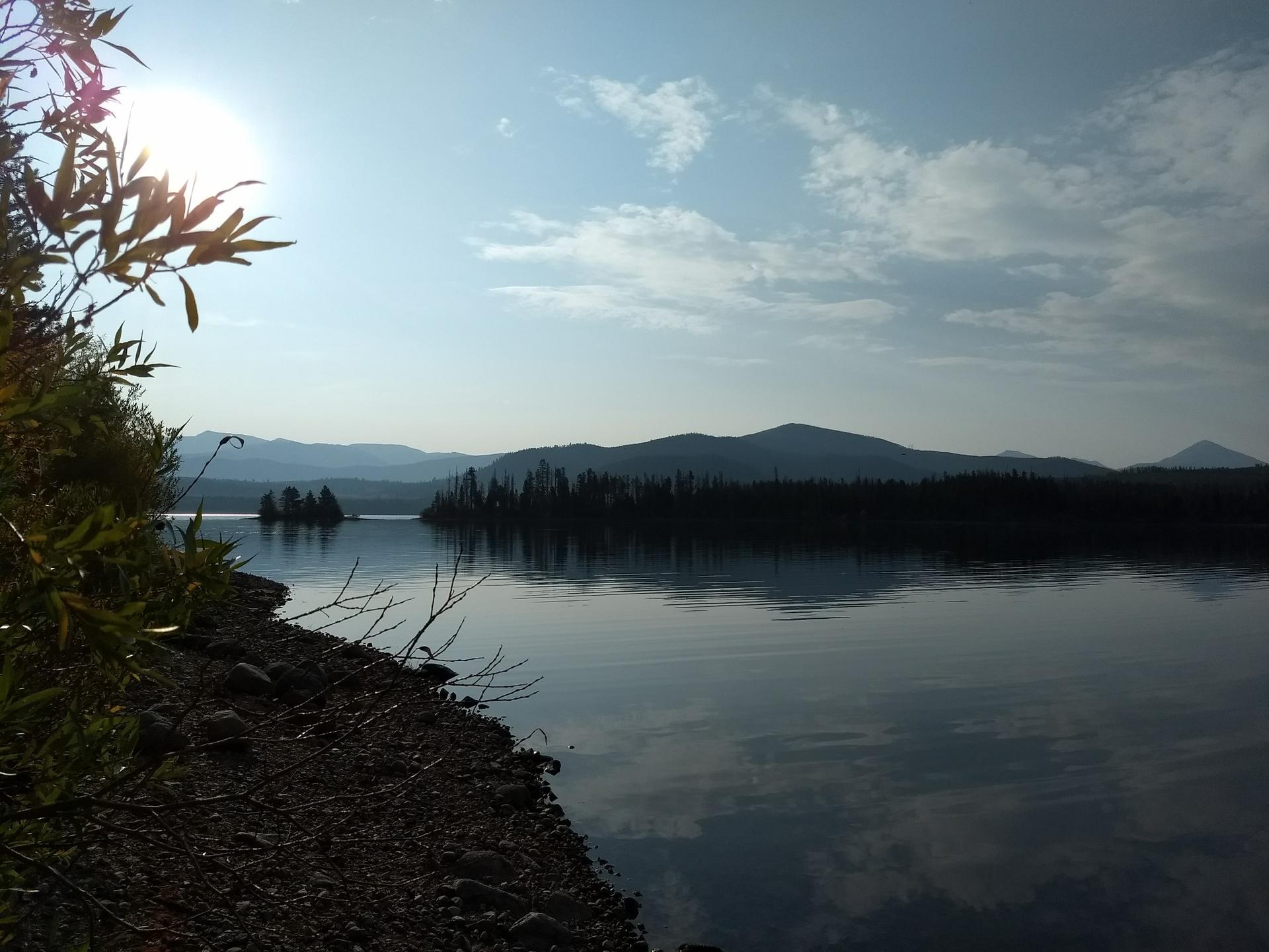 Sun shines over a calm lake with distant mountains and forested islands.