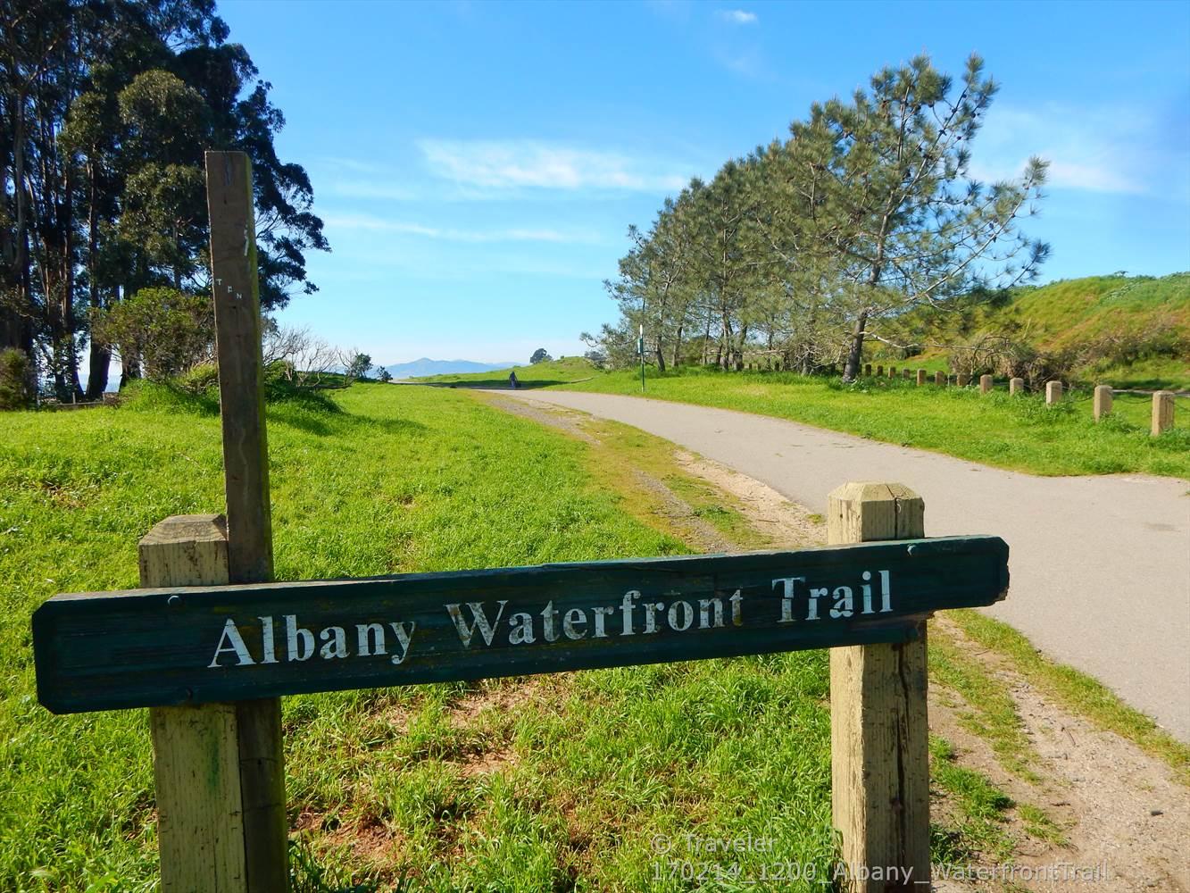 A wooden trail sign on a sunny day with a grassy path and trees.