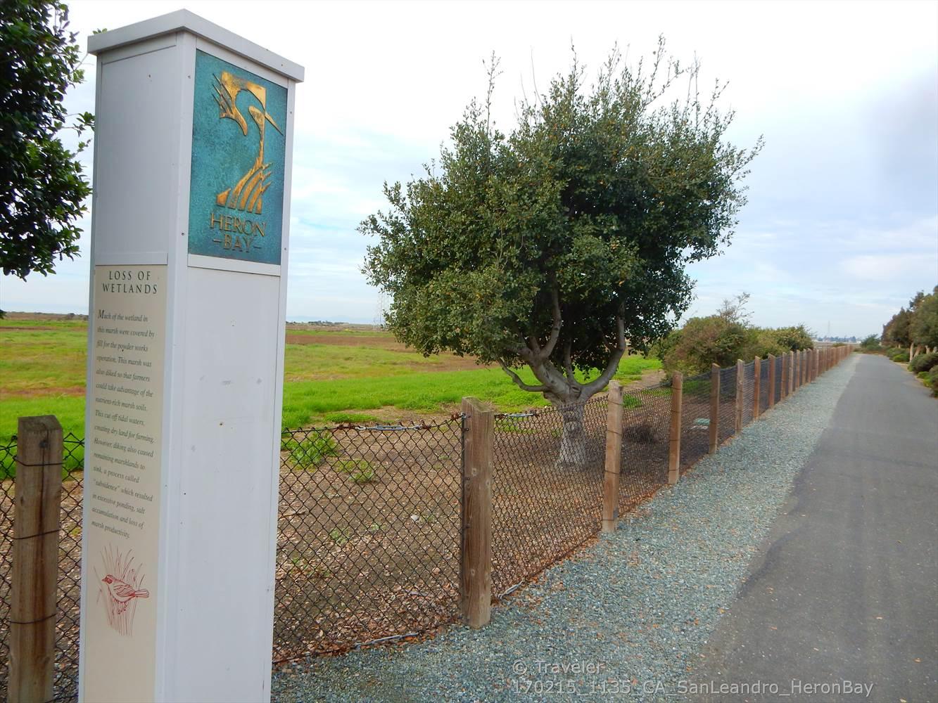 An interpretive sign stands beside a fenced path and green field.