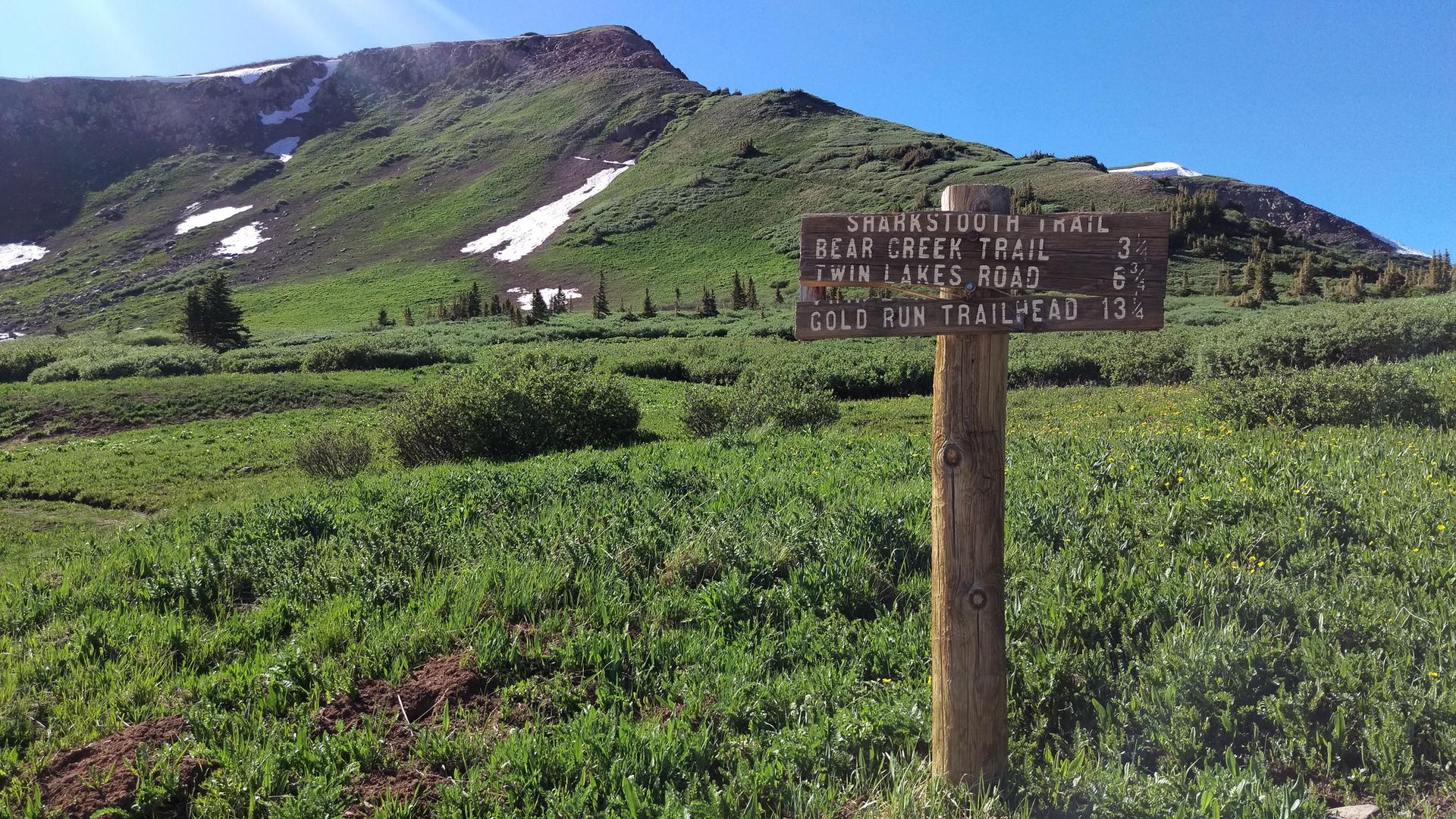 Wooden trail sign on a grassy hillside with a snow-dusted mountain behind.