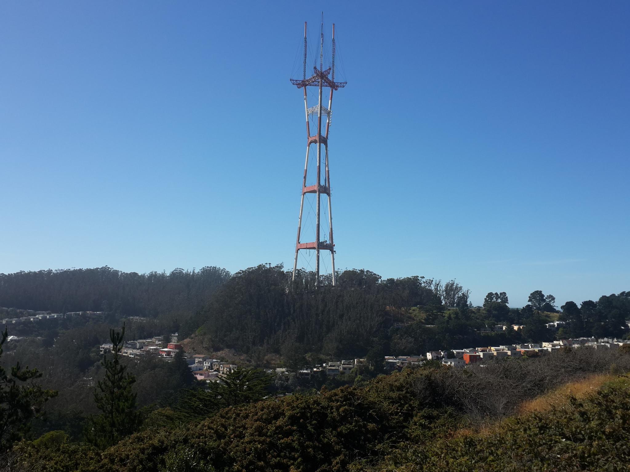 Tall communication tower on a hill, with distant city and trees.