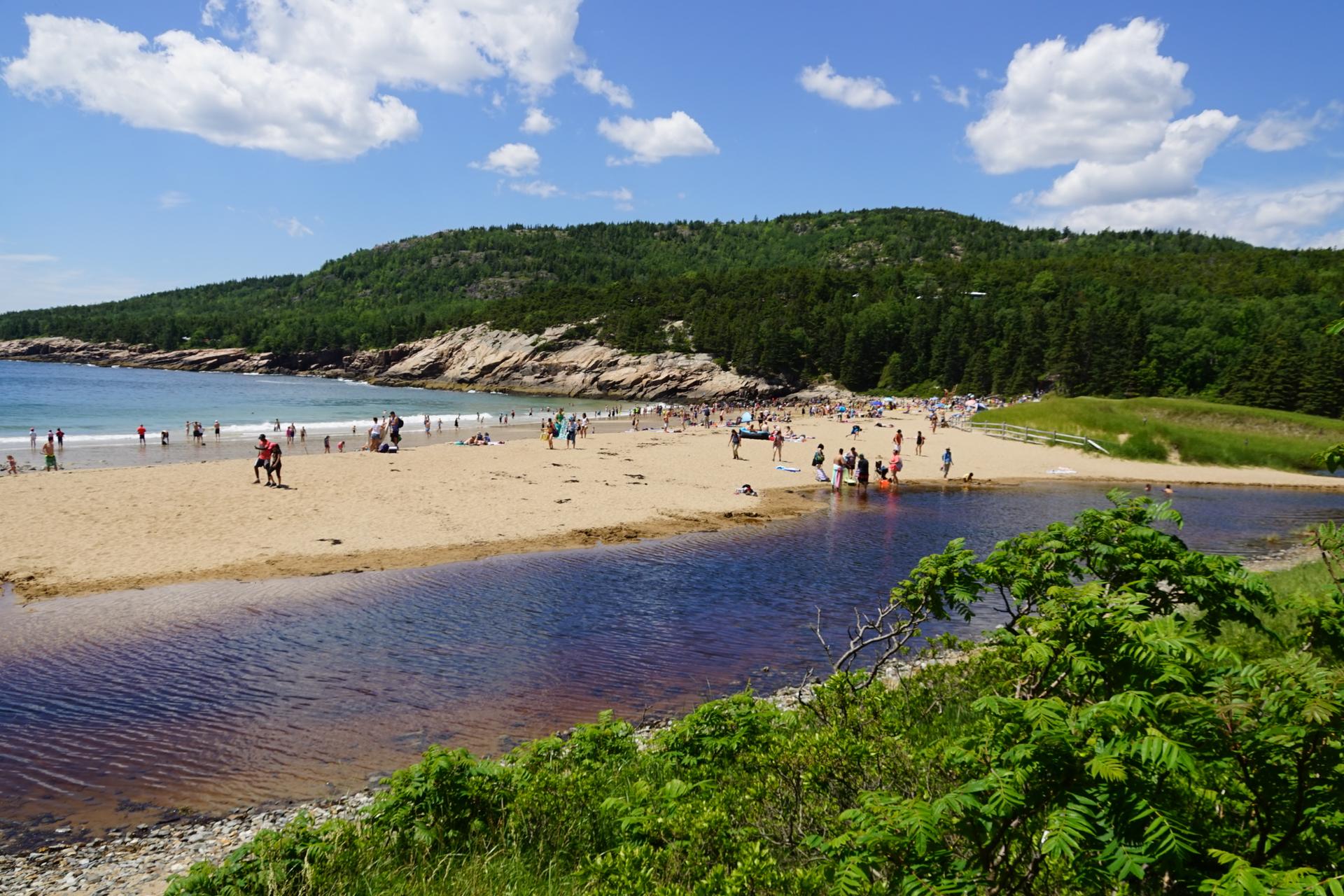 Sunny beach with many people, a river, and forested hills under blue sky.