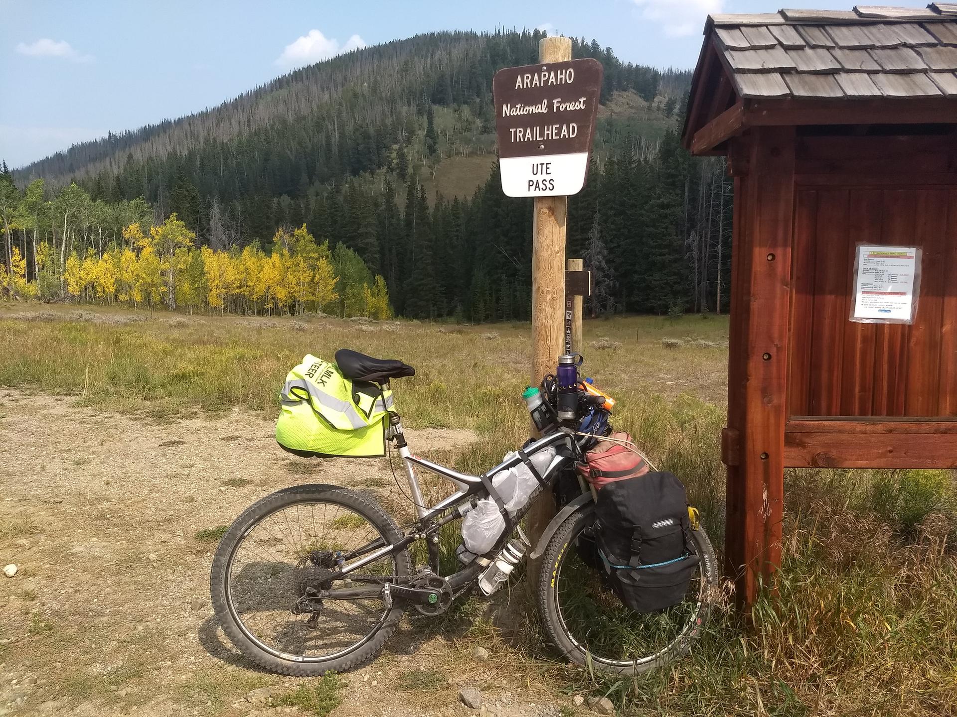 Loaded mountain bike at a trailhead sign, with autumn trees and mountains.