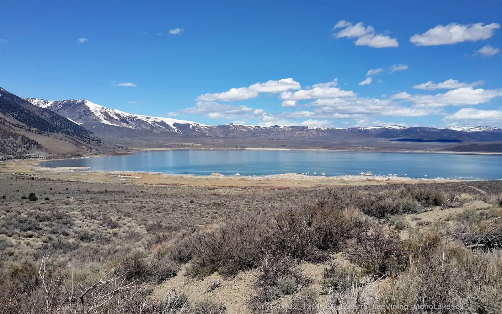 Wide shot of a large lake with snow-capped mountains under a bright blue sky.
