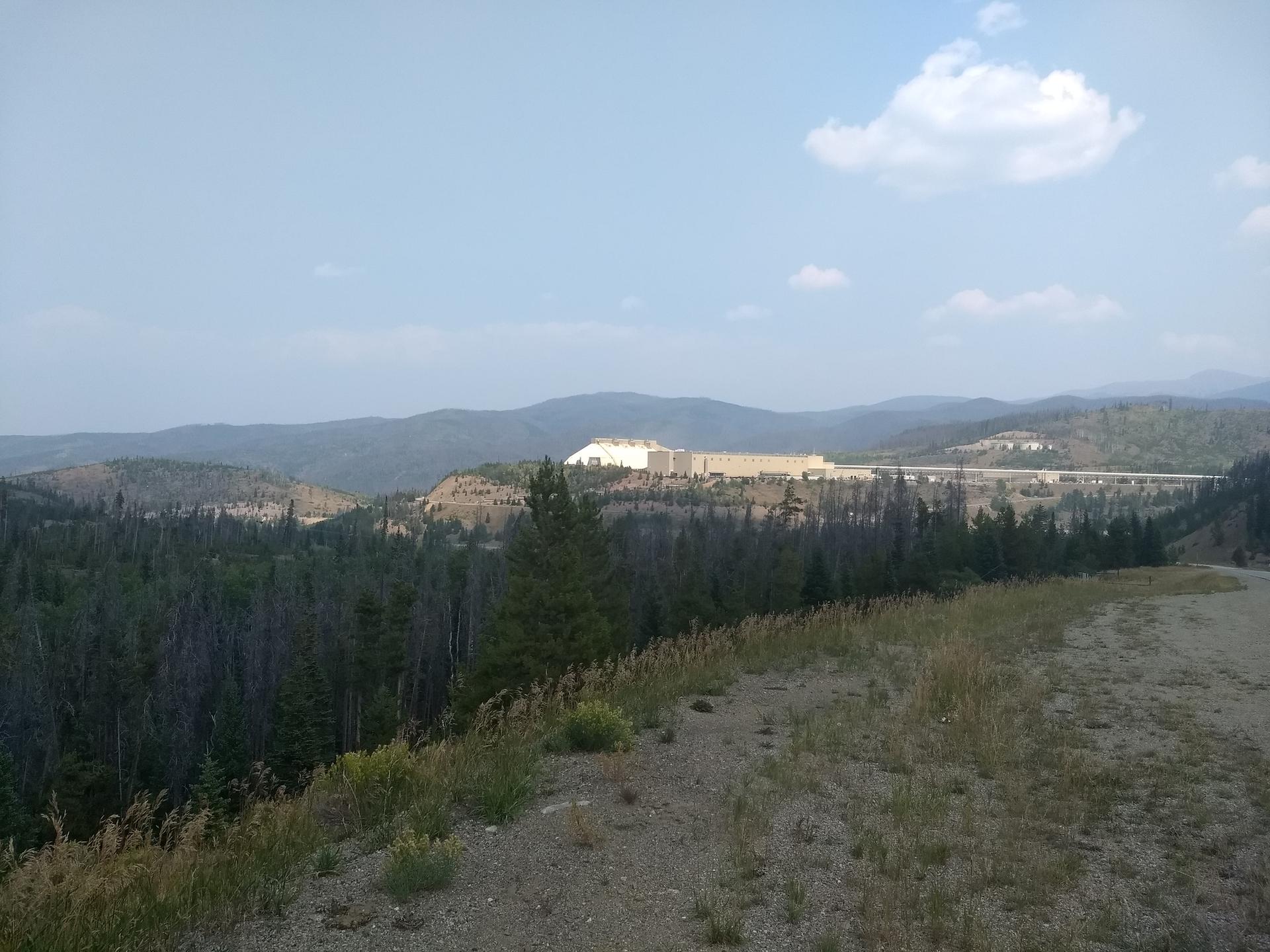 Distant industrial complex in a valley, surrounded by forested mountains, hazy sky.