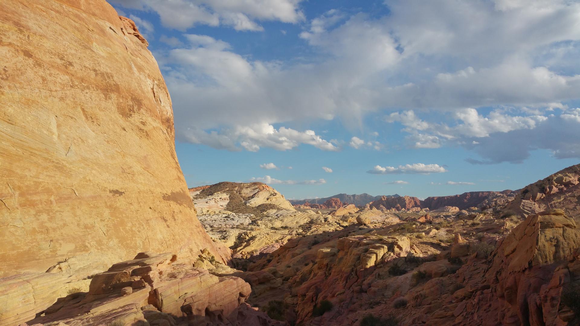 Panoramic desert view with towering red cliffs and distant mountains under a blue sky.