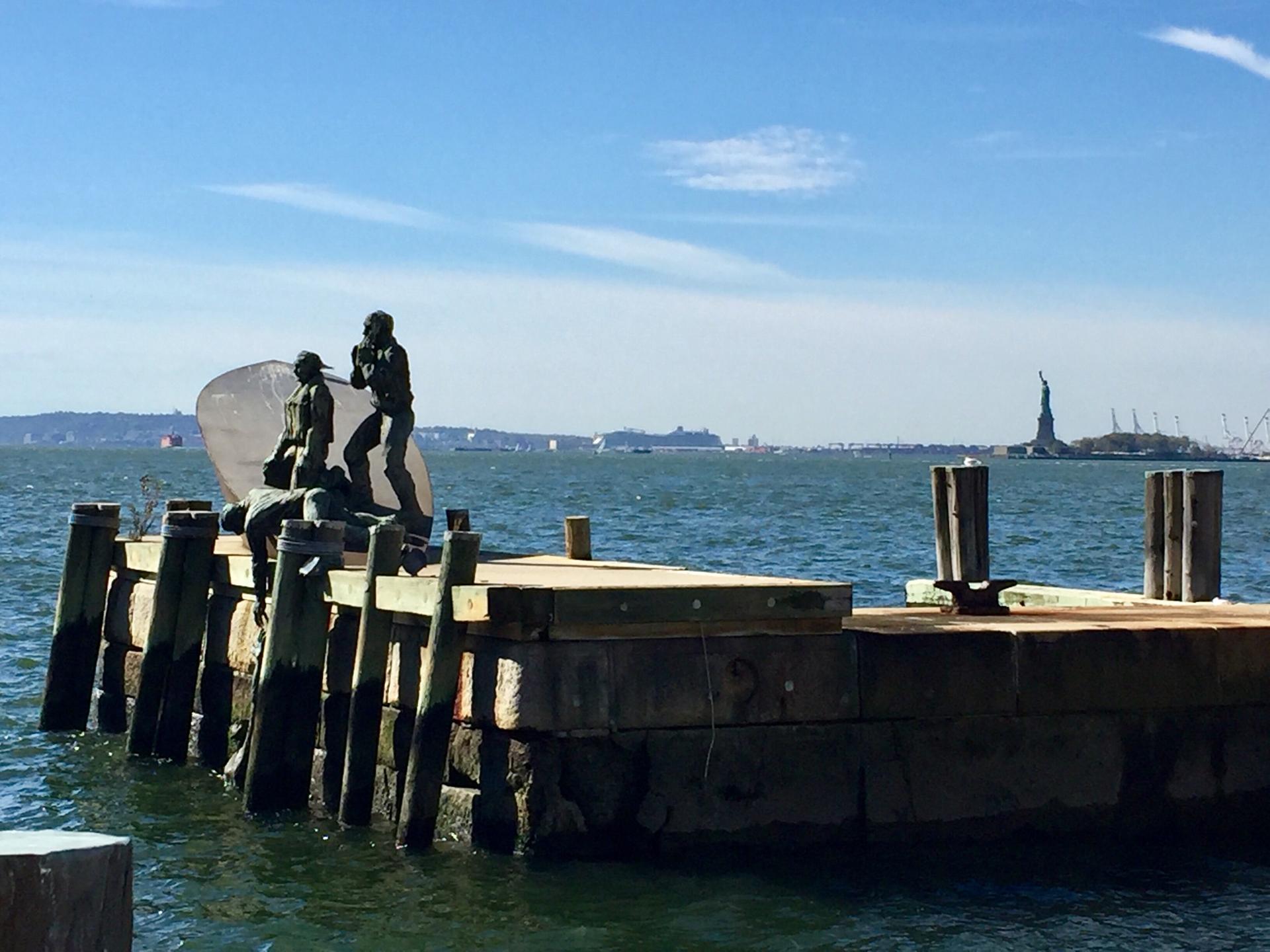 Statues on a wooden pier with a distant landmark across the water.