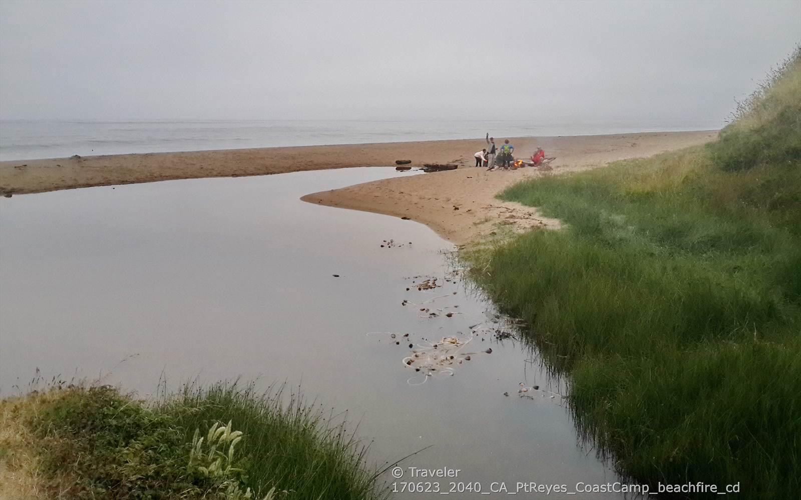 People gather on a sandy beach where a river meets the foggy ocean.
