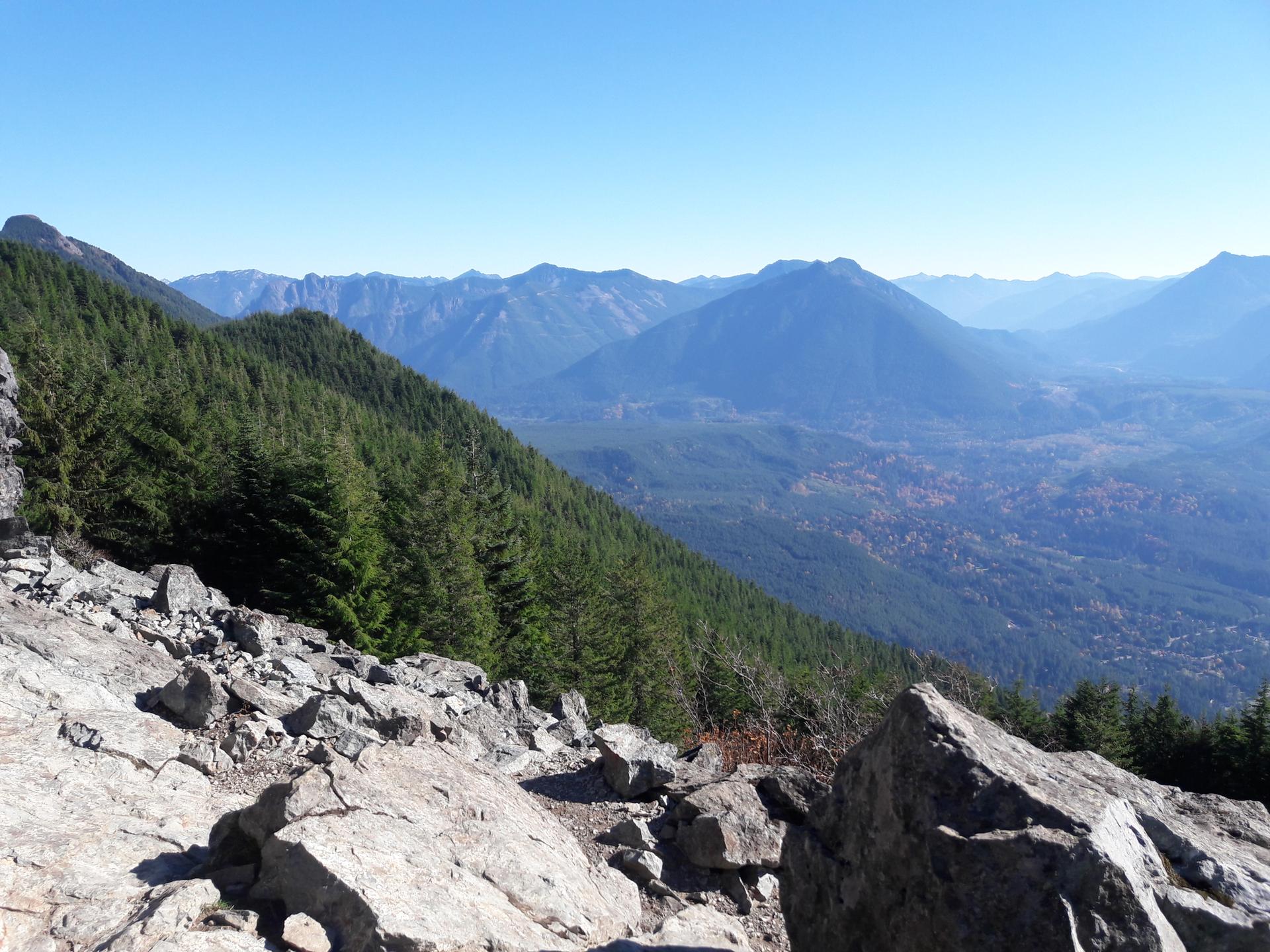 Rocky foreground with a forested mountain slope and hazy distant peaks under blue sky.