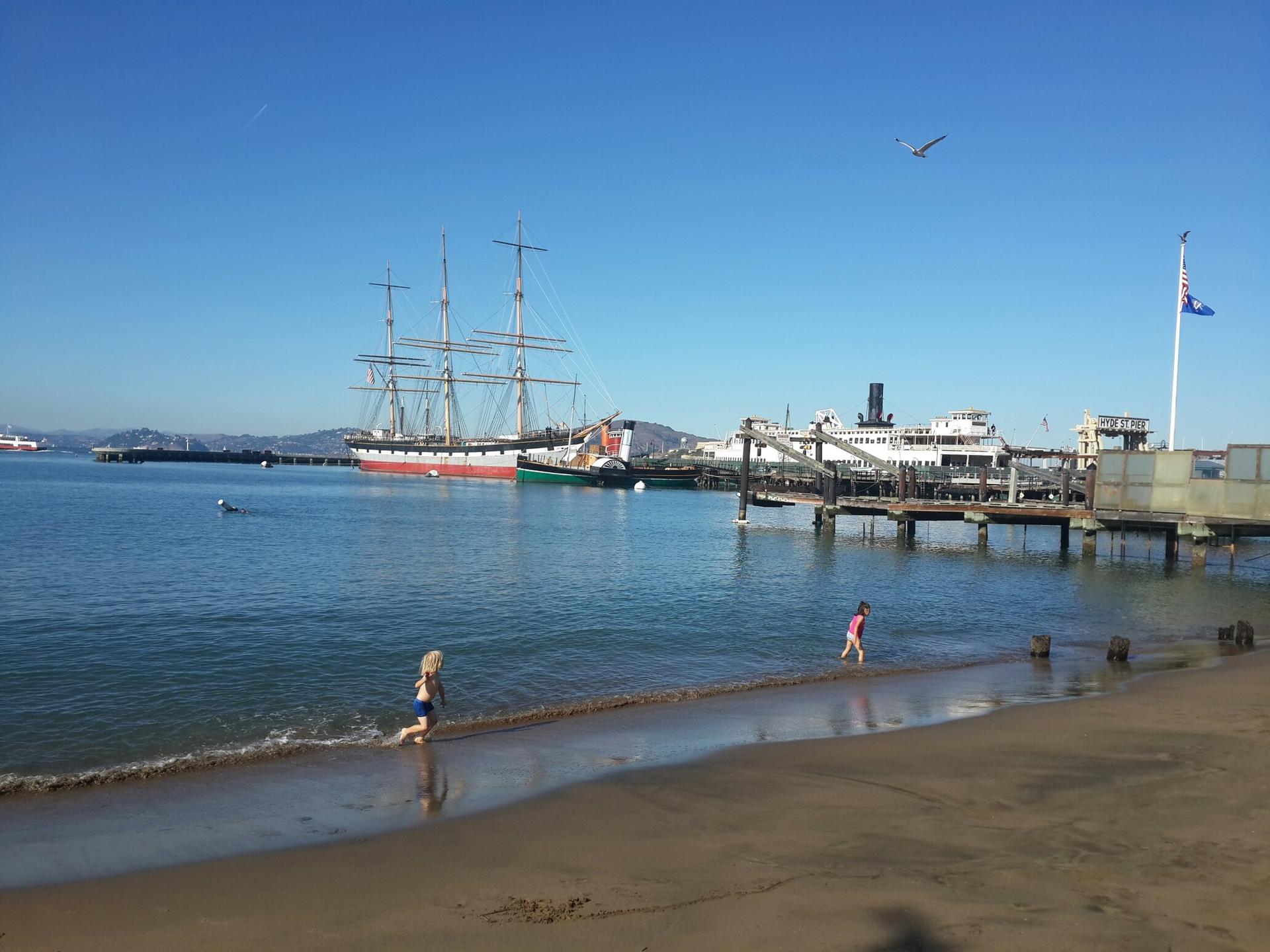 Children playing on a sandy beach, tall ships docked in a bay, sunny.