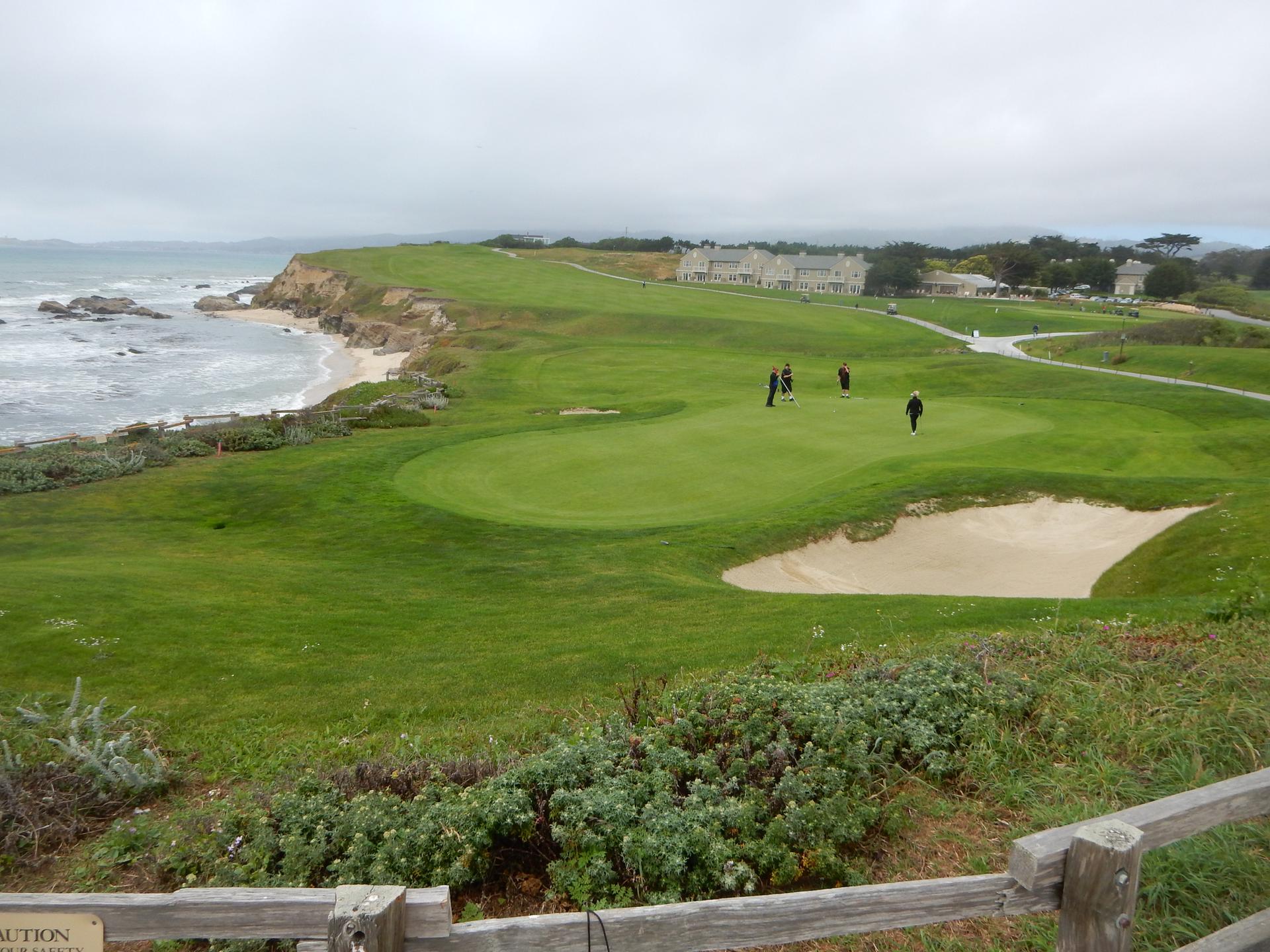 Golfers on a green with a sand trap, ocean, and distant building under cloudy sky.