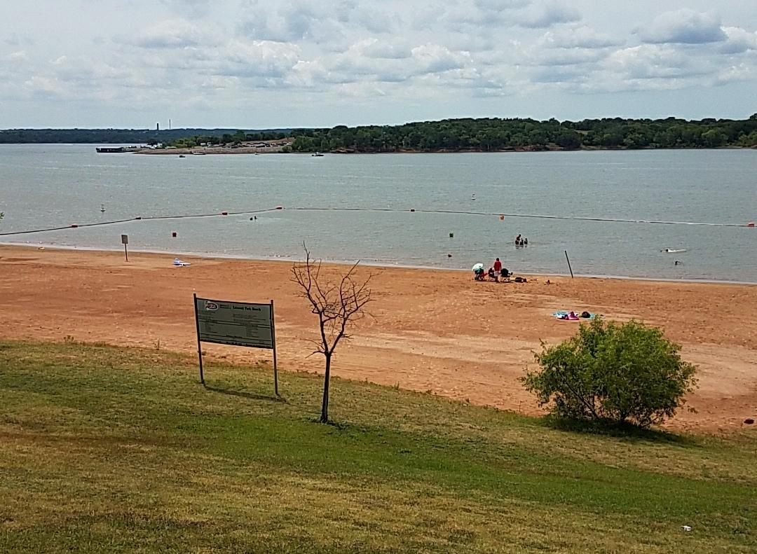 People swim in a lake next to a sandy beach under an overcast sky.