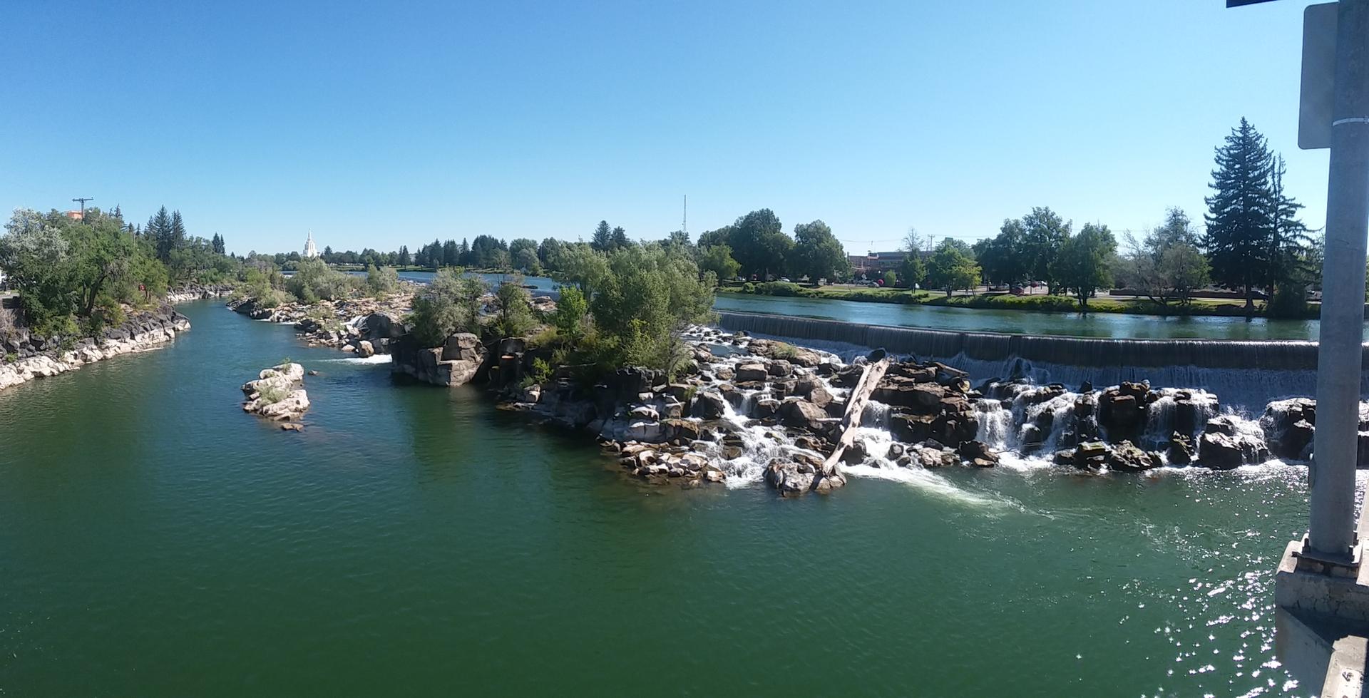 Wide river with rocky islands and a waterfall under a clear blue sky.