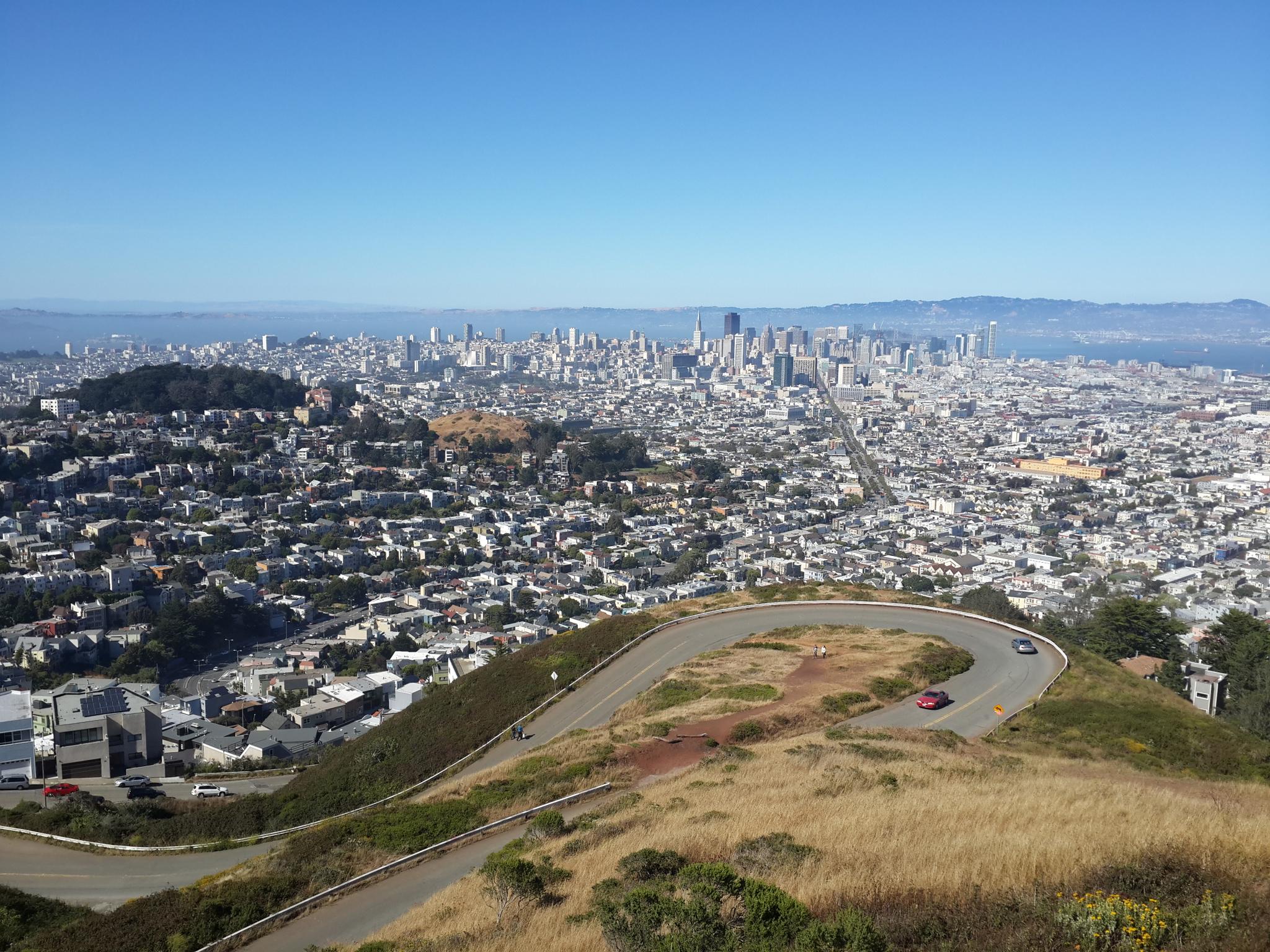 Sunny city panorama from a hill, with a winding road and distant skyline.
