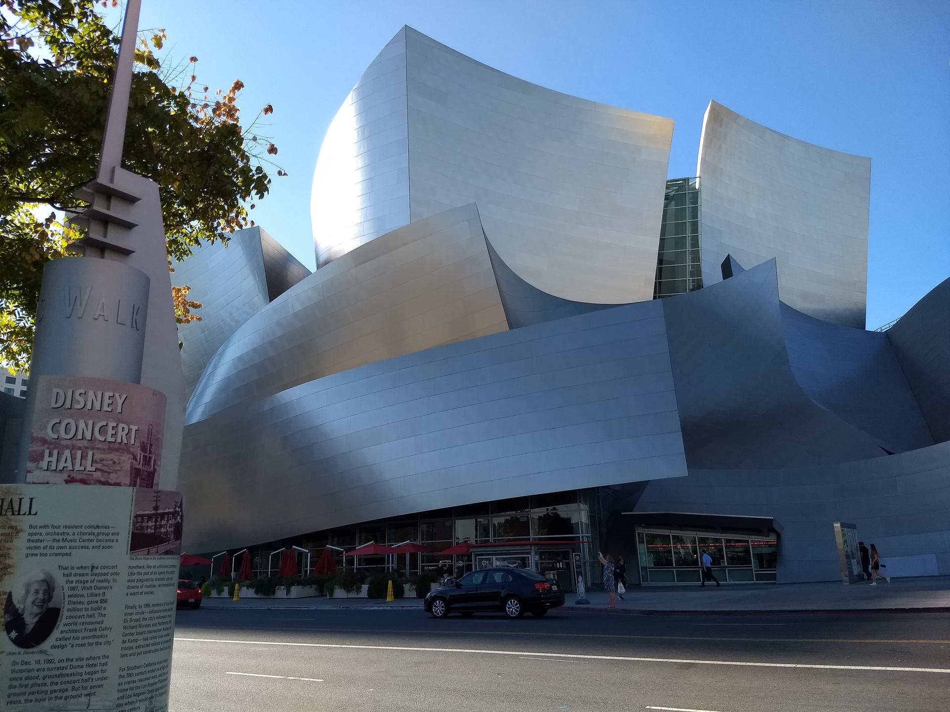 Modern building with curved metallic panels, a car, and people on a sunny day.