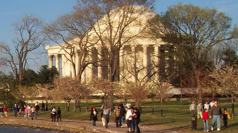 Large domed building surrounded by trees and many people walking, sunny.