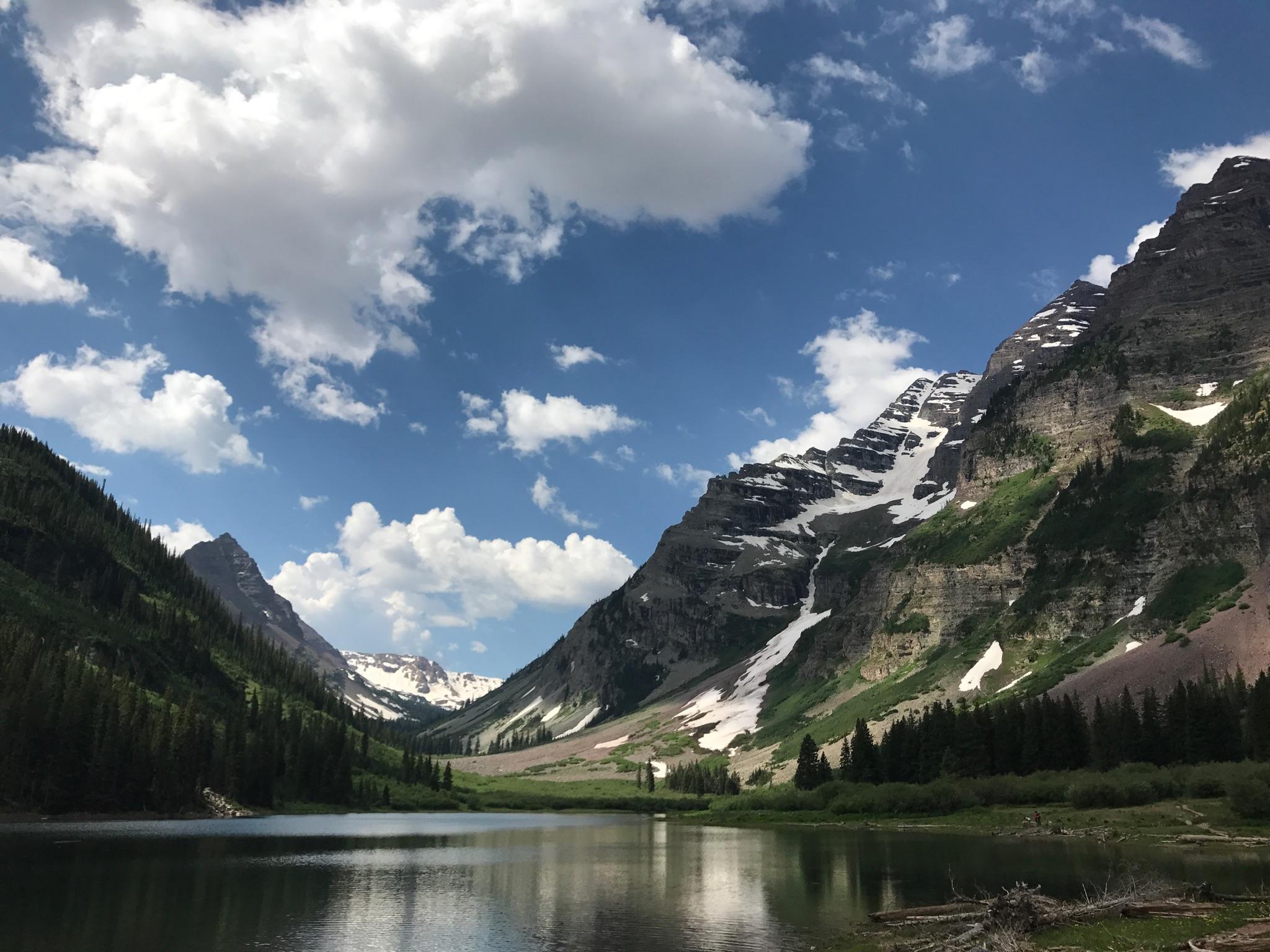 Calm lake in a valley with snow-capped mountains under a cloudy blue sky.