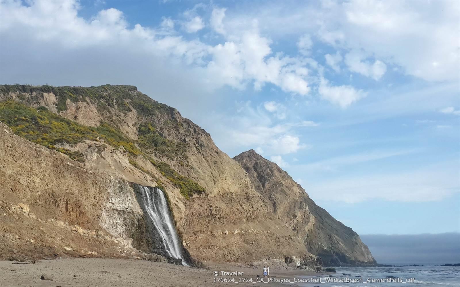 Waterfall cascading down a large, rocky cliff onto a sandy beach under a cloudy sky.