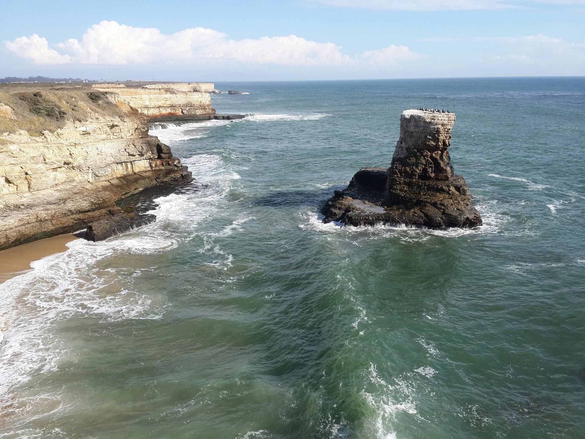 Sunny view of a sea stack, ocean waves, and rugged cliffs.