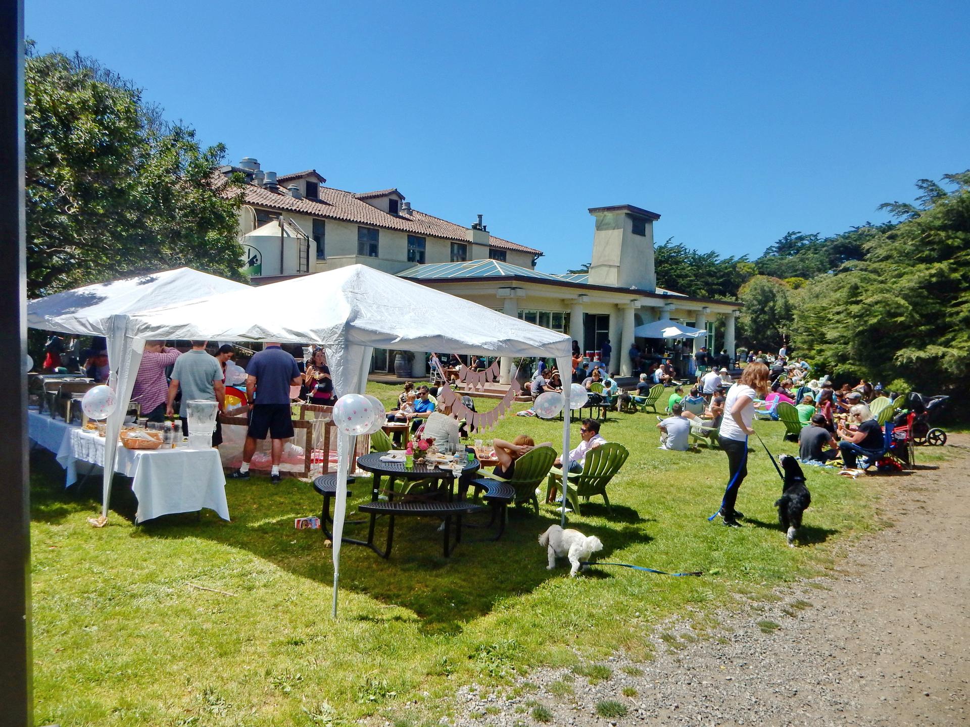 Sunny outdoor gathering with white tents, people, and a large building.