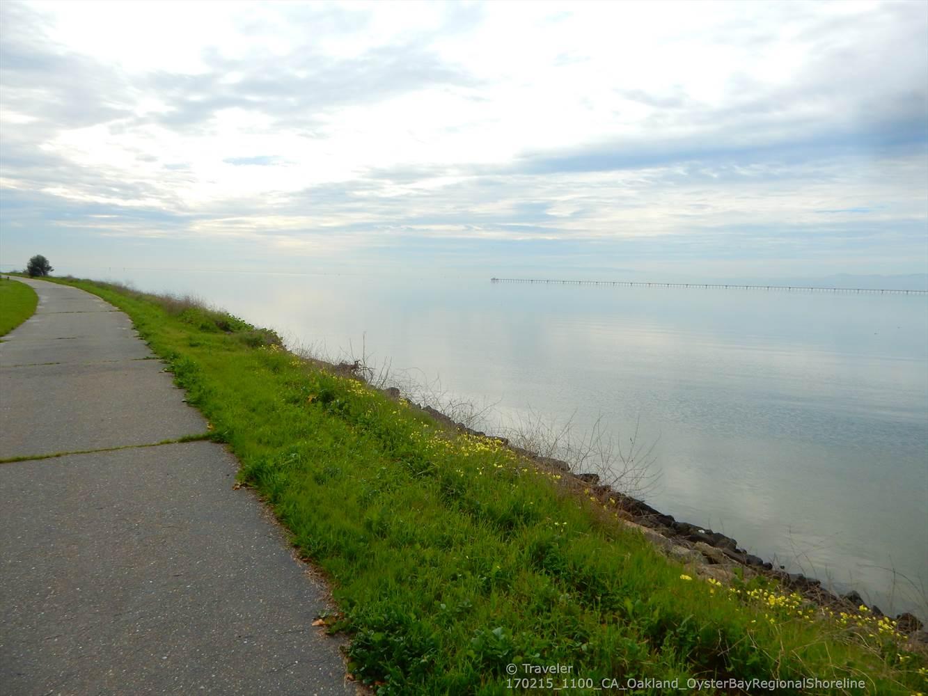 A paved path runs alongside calm water under a cloudy sky.