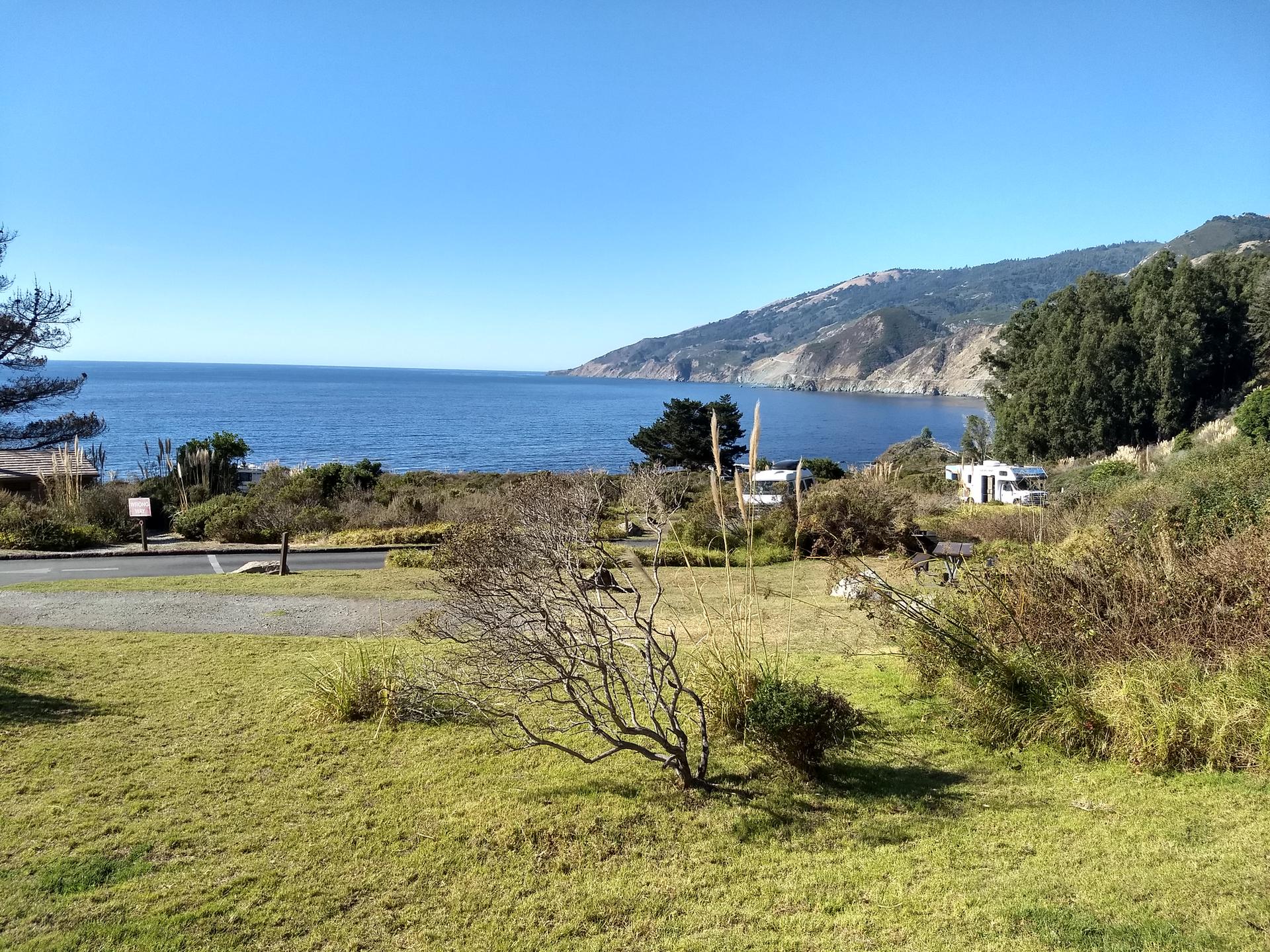 Sunny coastal view with blue ocean, distant mountains, and RVs near shore.