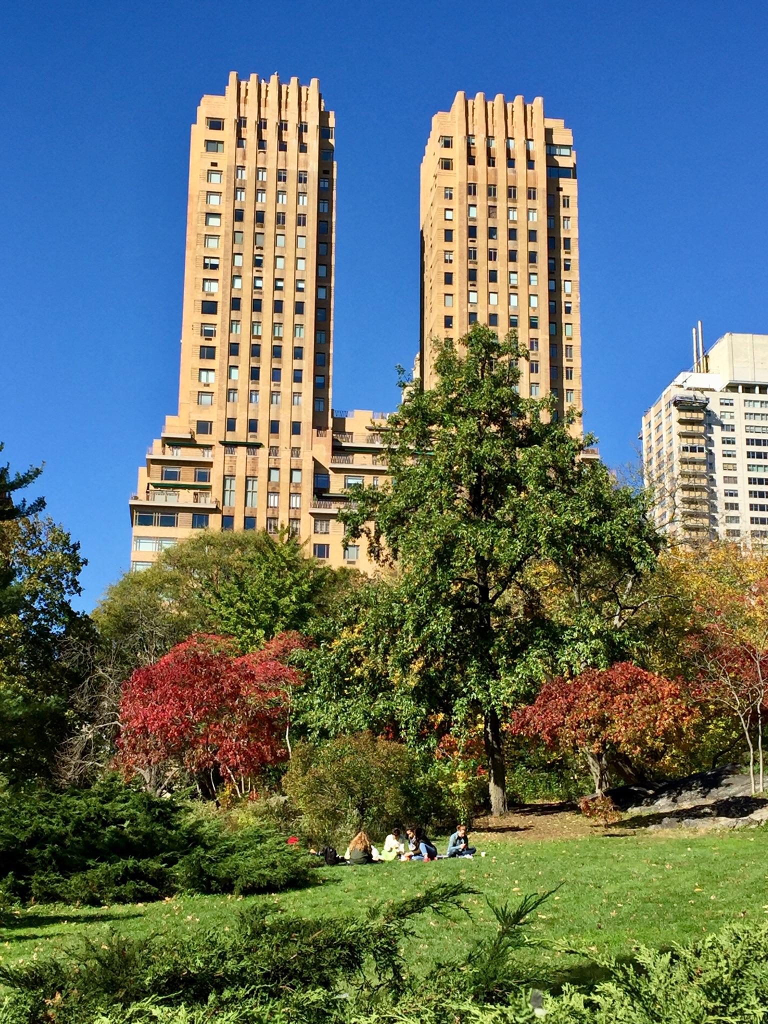 Tall buildings behind colorful autumn trees, people relaxing on green grass.