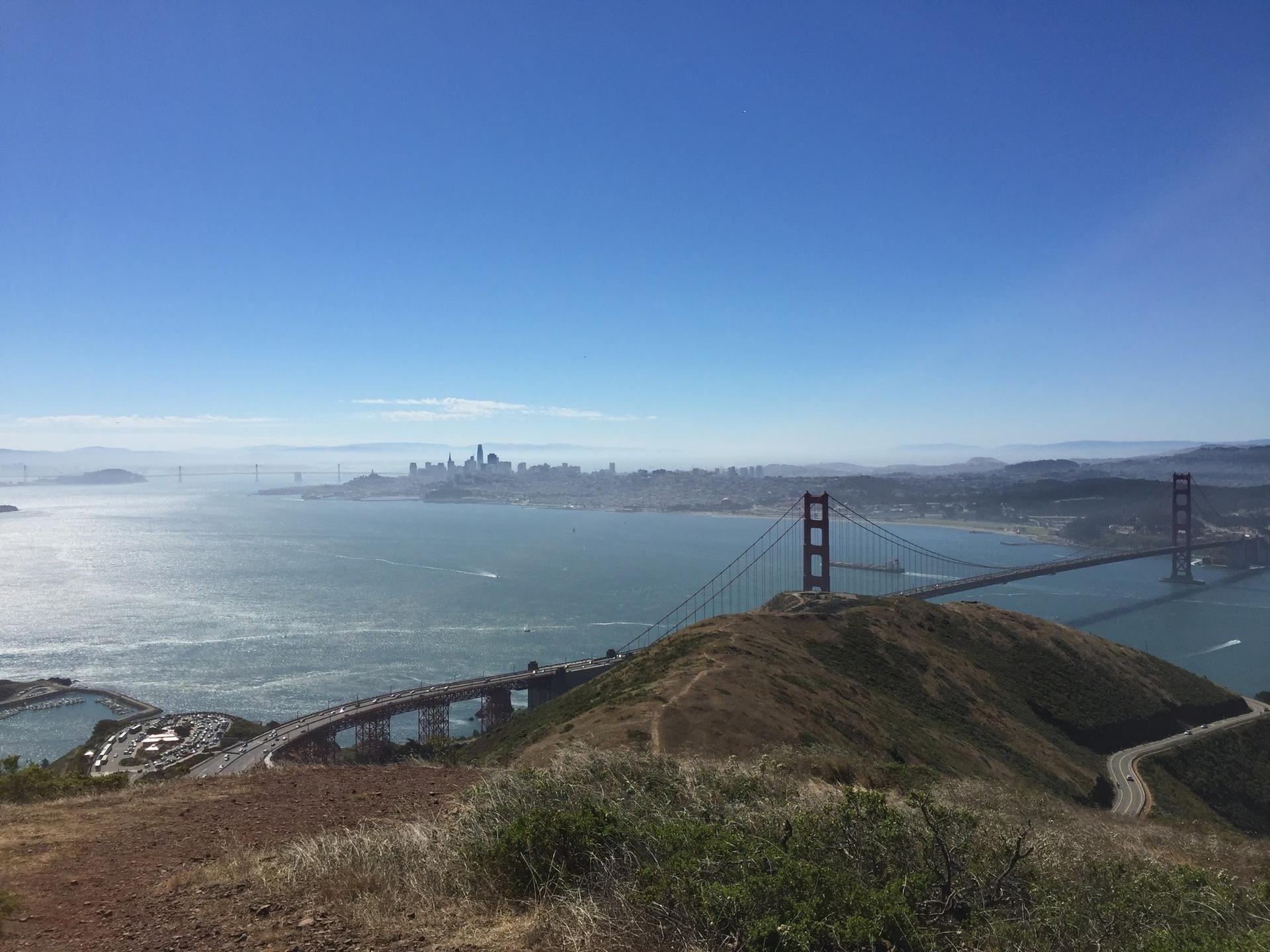 Panoramic view of a large red suspension bridge, bay, and distant city.