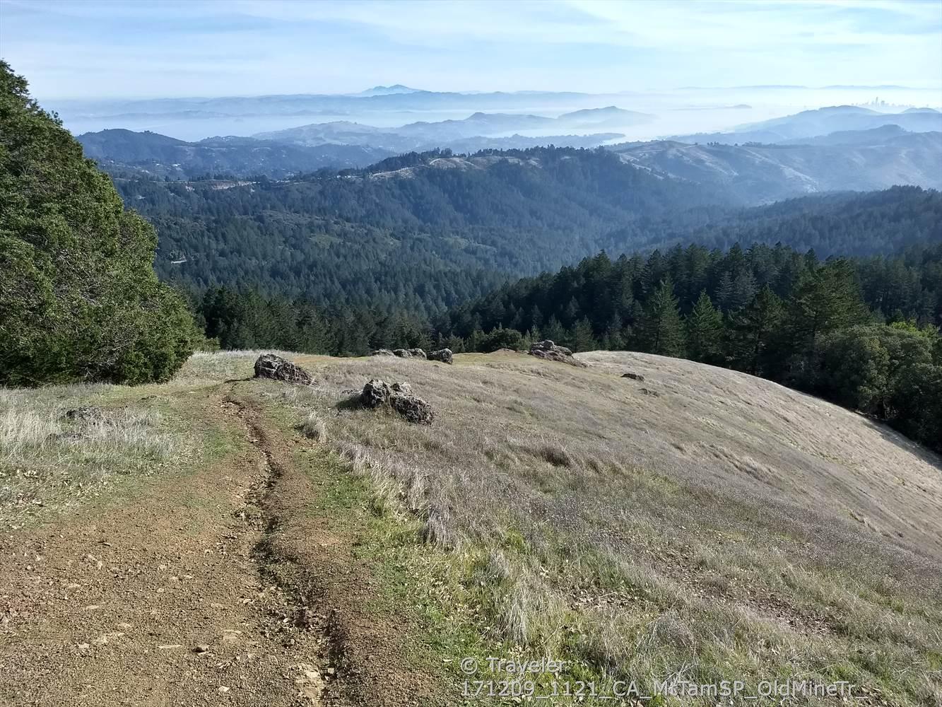 Dirt path on a grassy hilltop overlooks distant hazy mountains and forests.