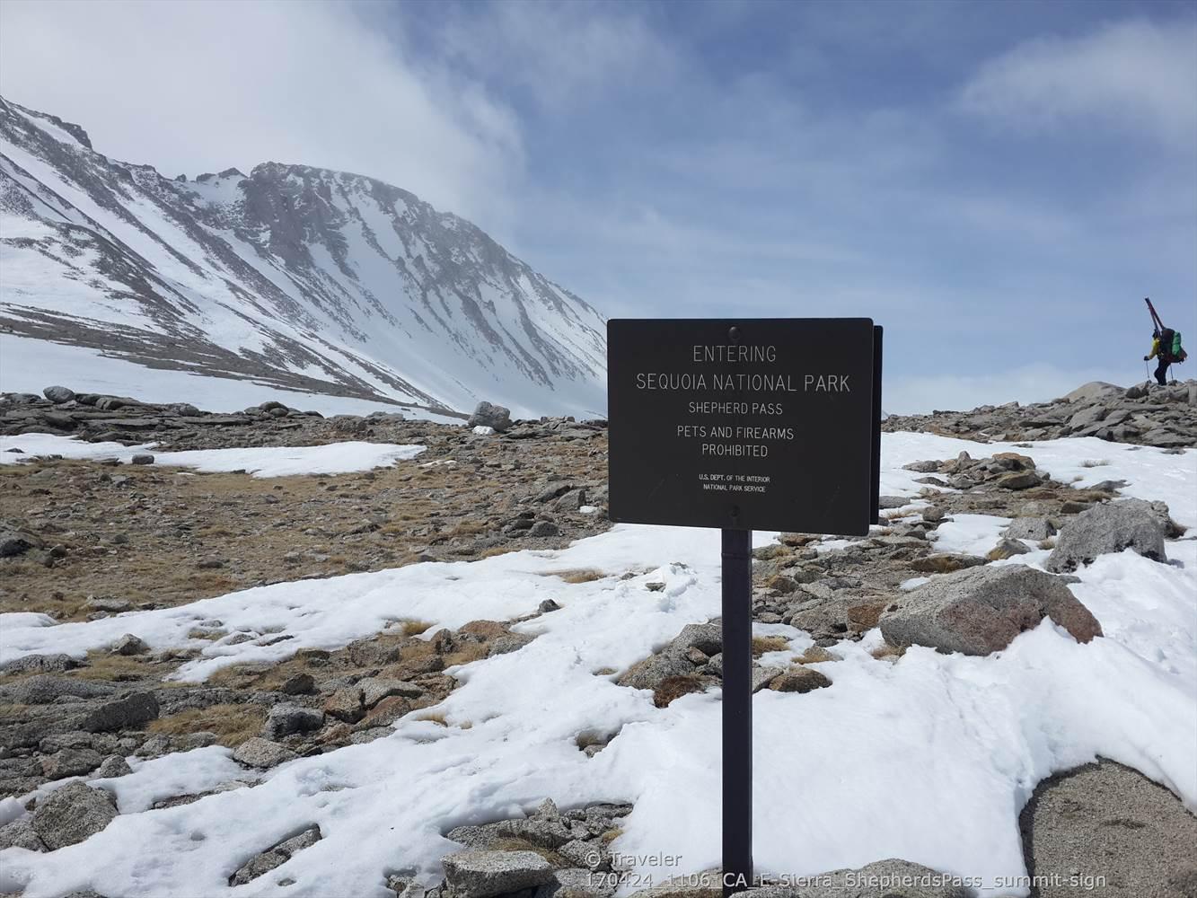 A sign in a snowy, rocky landscape with a distant snow-capped mountain and hiker.