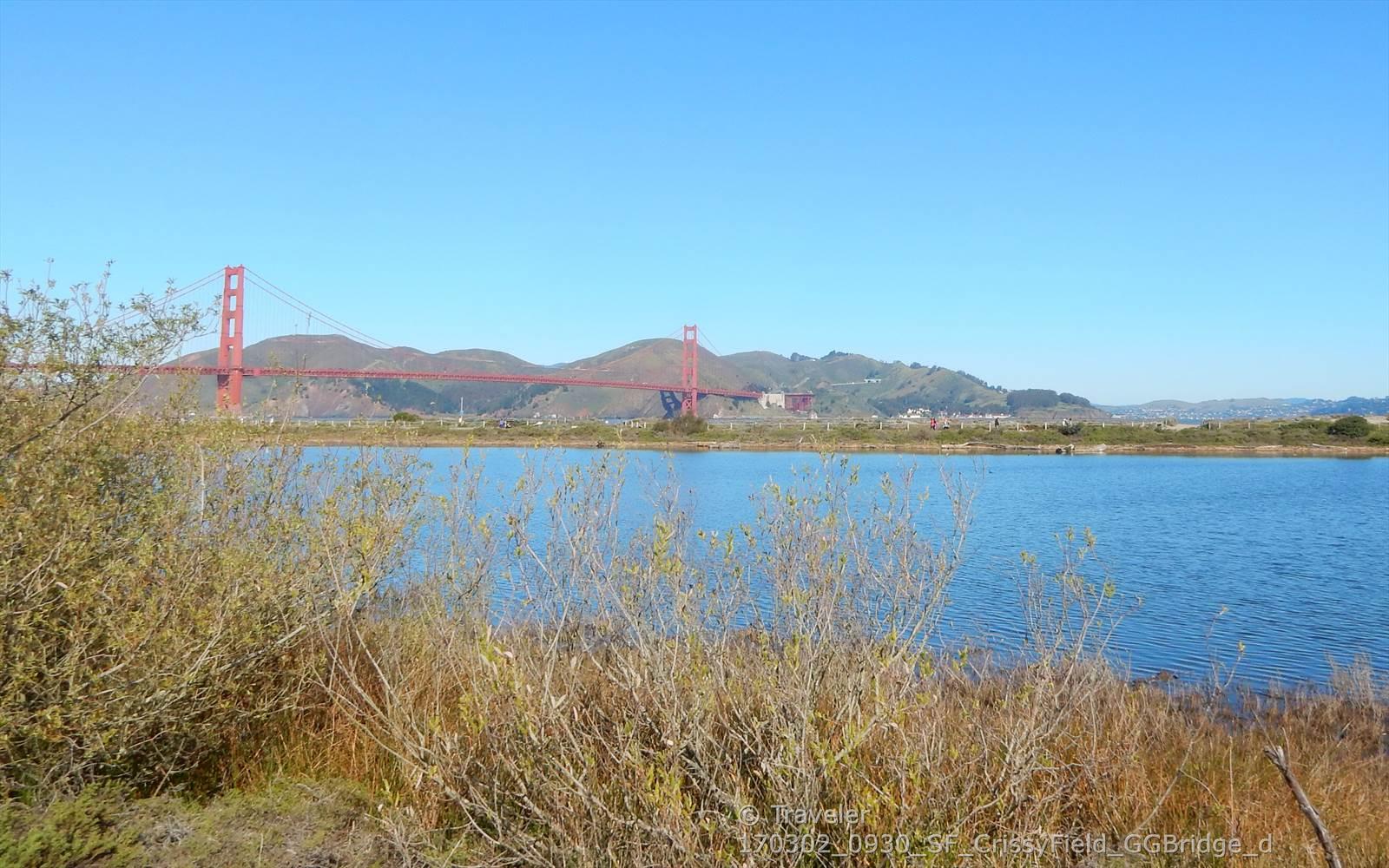 A red suspension bridge spans water with hills under a clear blue sky.