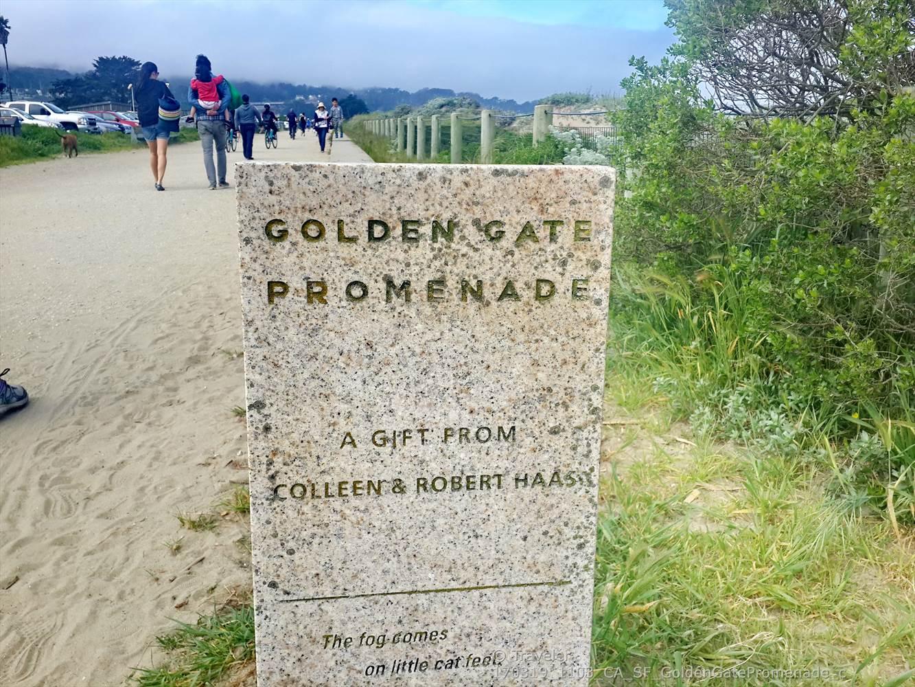 Stone sign on a sandy path next to green bushes, people walking.
