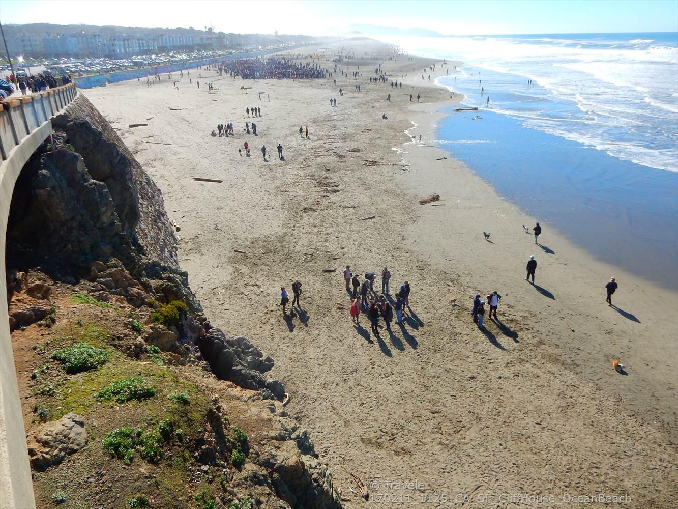 Crowded sandy beach with ocean waves and a concrete wall on a sunny day.