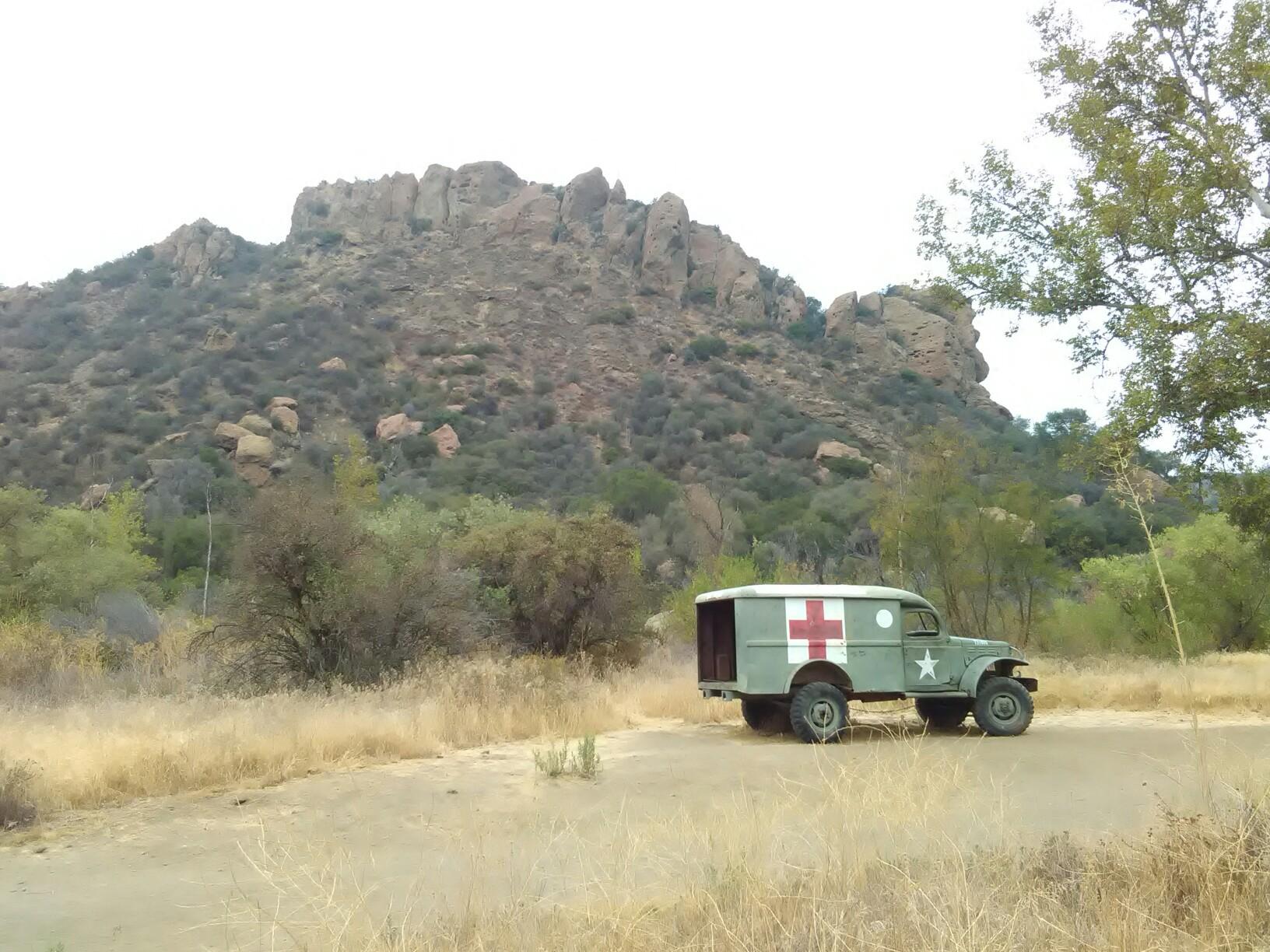 Vintage military ambulance parked on a dirt road near rocky hills.