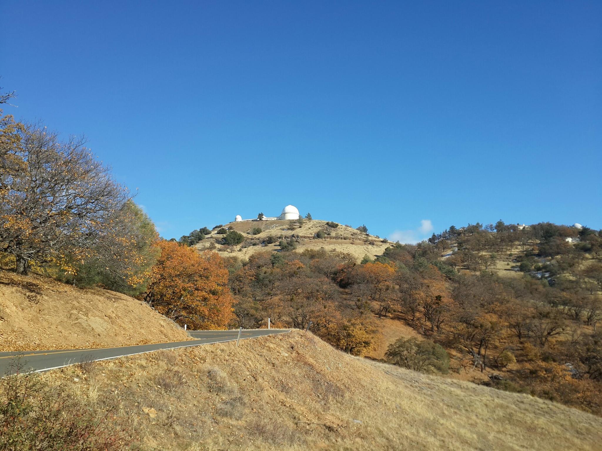 Sunny view of a winding road leading to observatories on a distant, tree-covered hill.