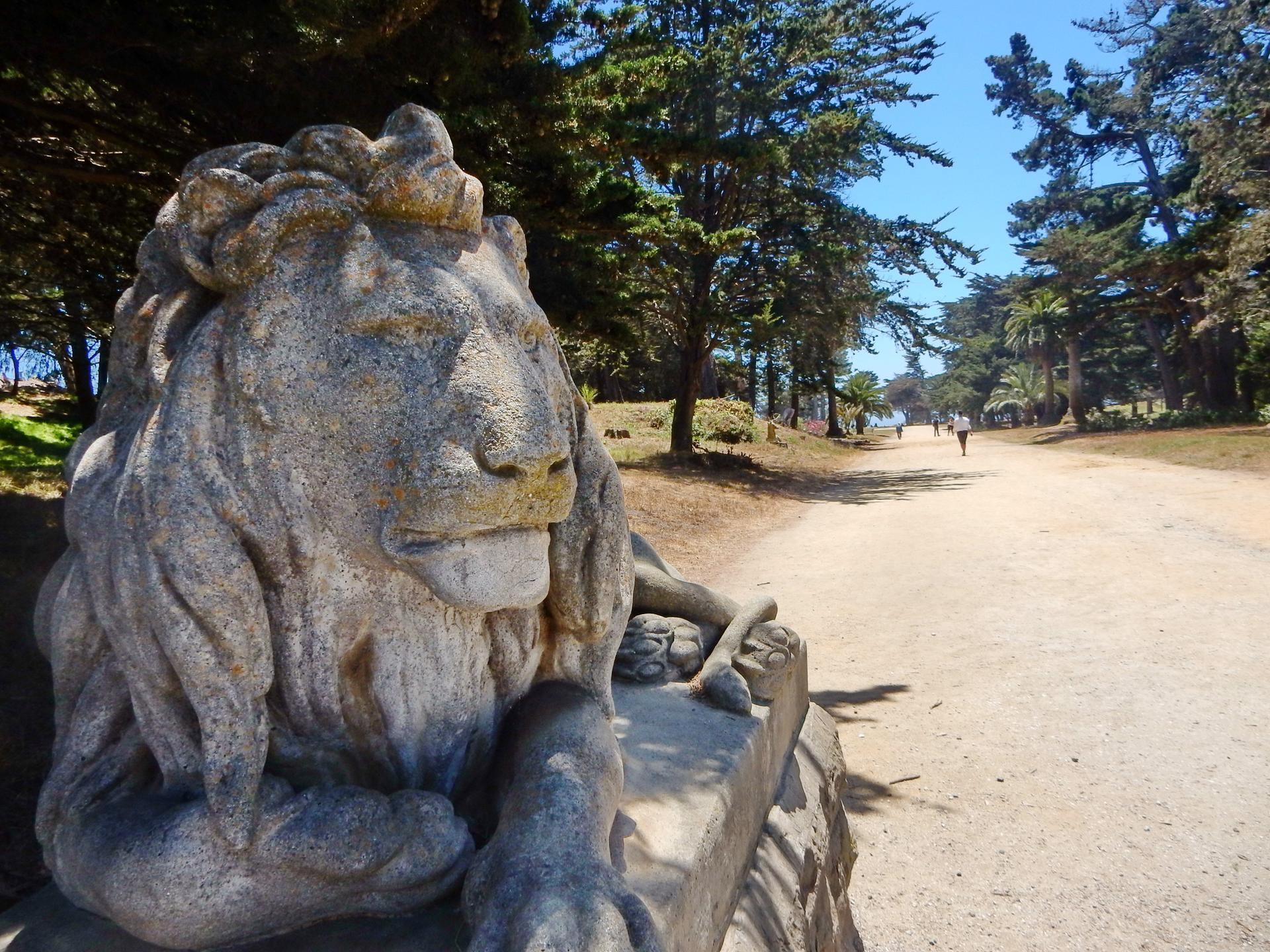 Stone lion statue beside a sunny dirt path lined with tall trees.
