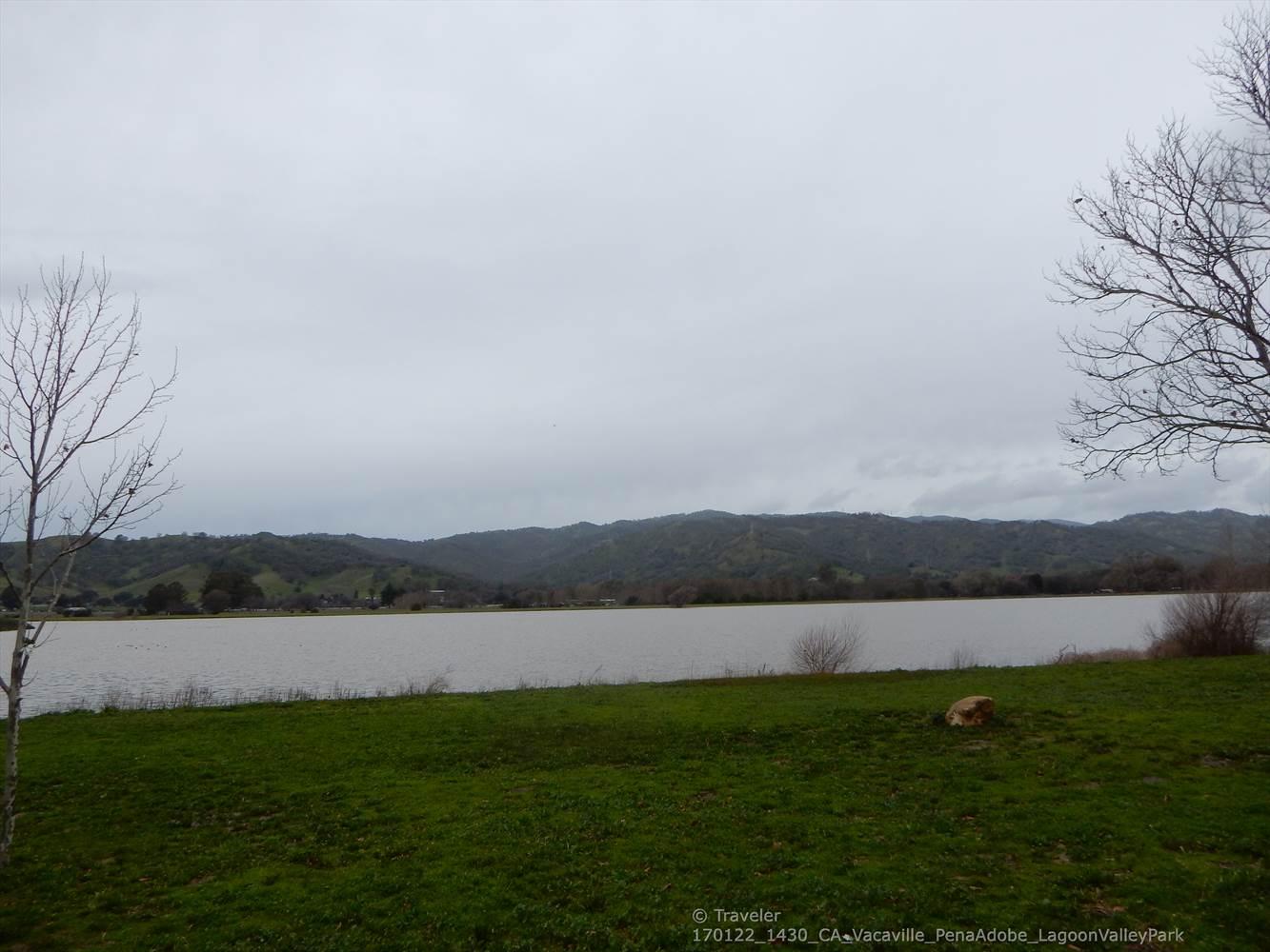 Overcast sky above a calm lake, green hills, and bare trees.