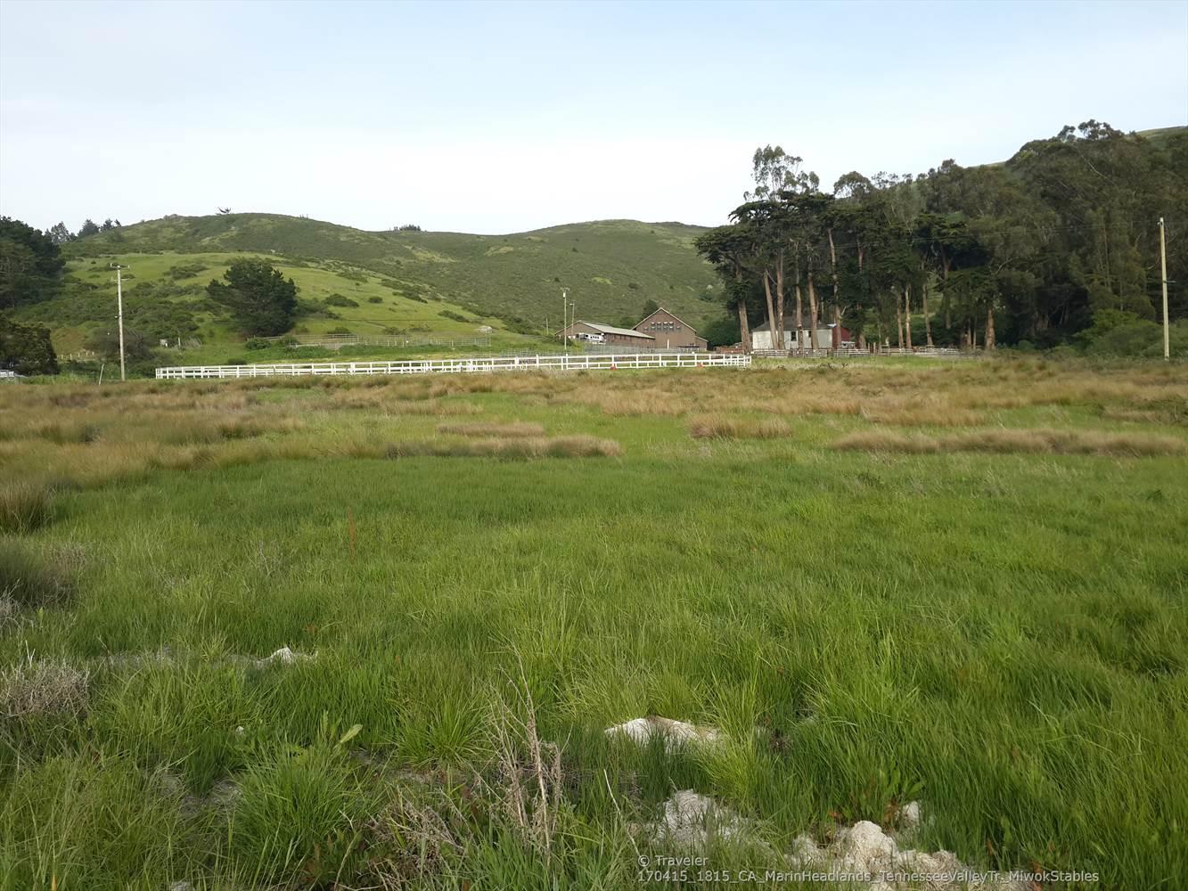 A wide green field with distant buildings, trees, and rolling hills.